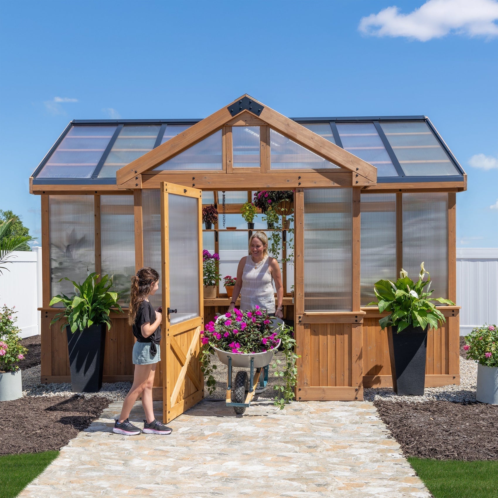 Savannah cedar greenhouse with polycarbonate panels and peaked roof, woman with wheelbarrow exiting the door.
