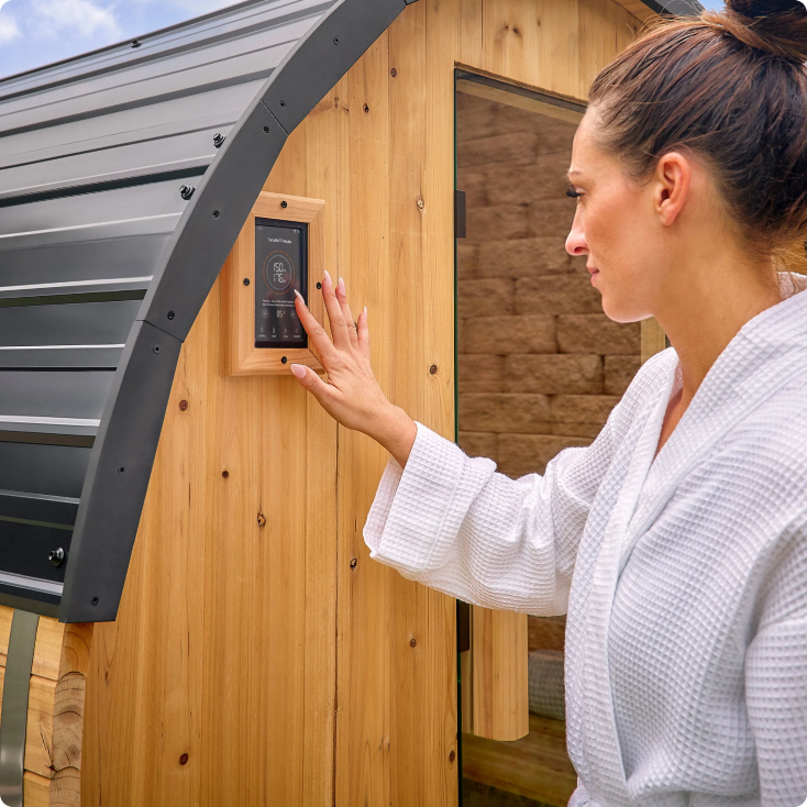 Woman in a white robe interacting with a digital display on a wooden outdoor sauna.