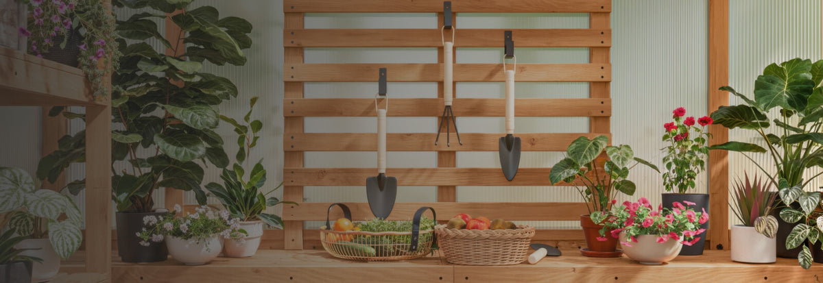 A wooden slat wall-mounted tool rack holding two garden trowels, styled in a garden room with potted plants, baskets of fruit, and a wooden bench.
