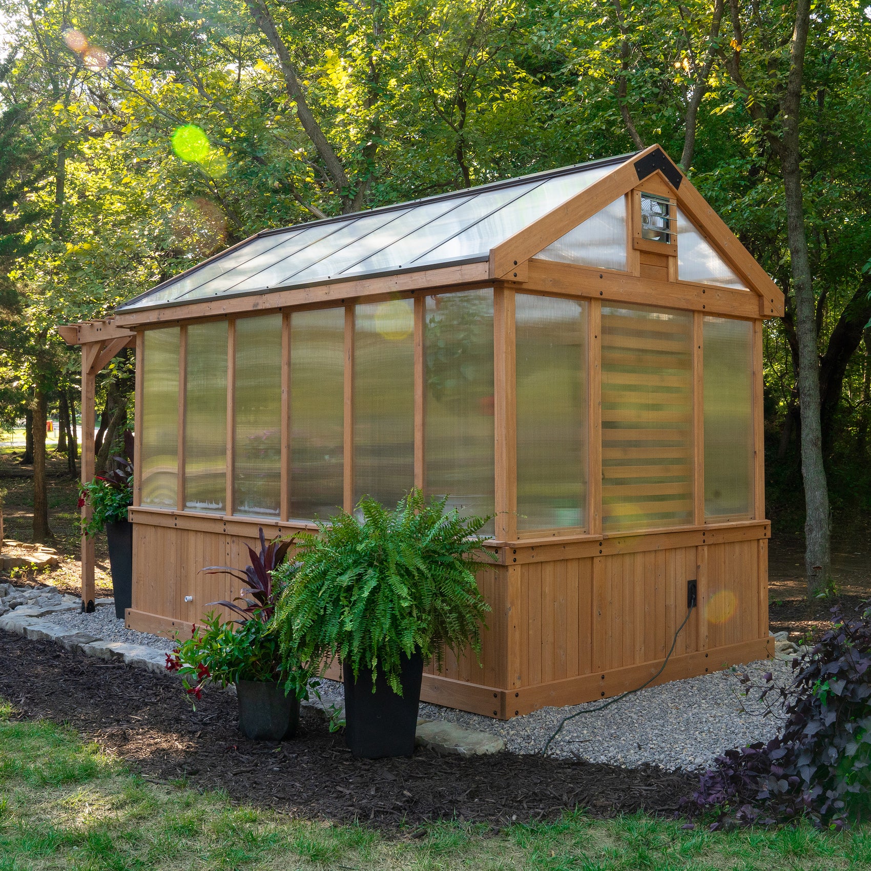 Bellerose cedar-frame garden greenhouse with polycarbonate panels set among lush trees with potted ferns outside.