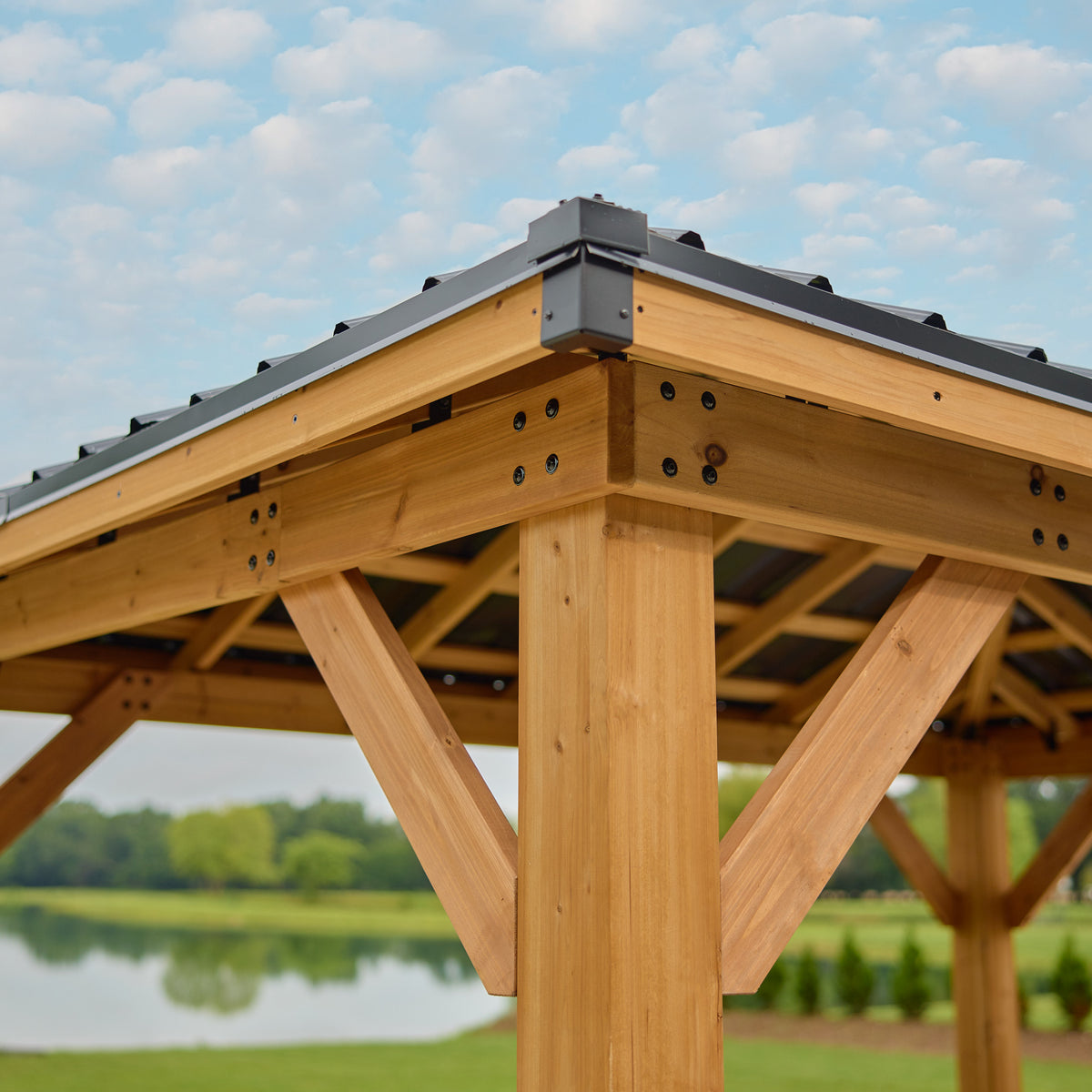 Black steel hip roof joining at the corner peak above cedar beams and posts of an outdoor gazebo.