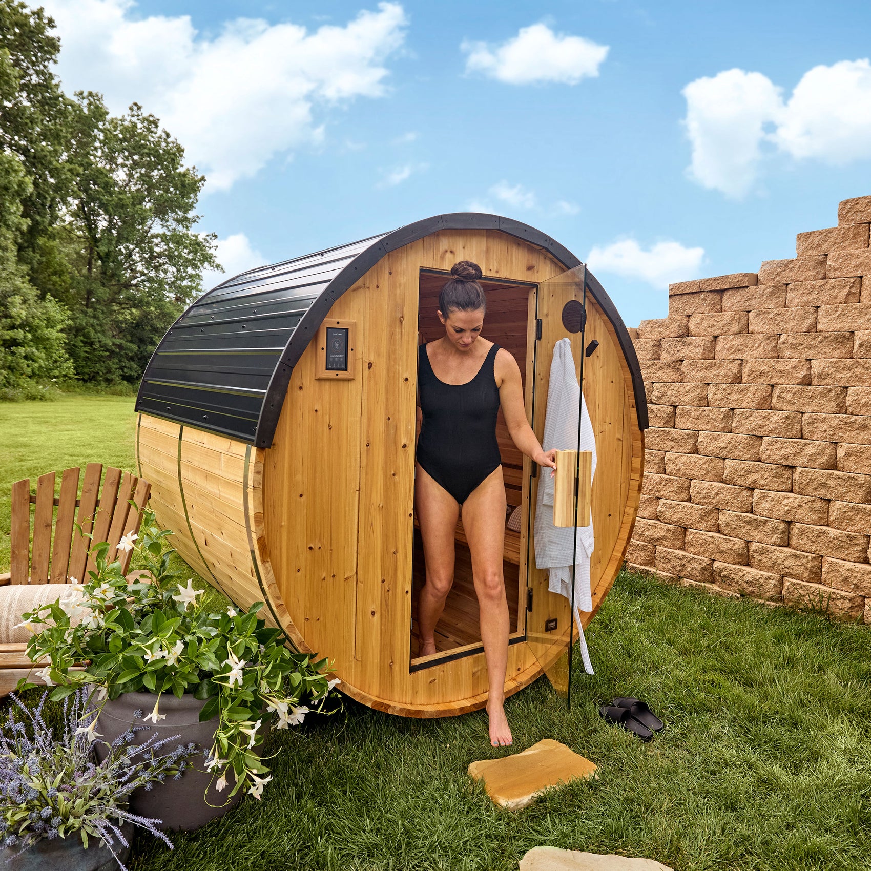 Woman exiting Paxton cedar barrel sauna with white towel in sunny backyard