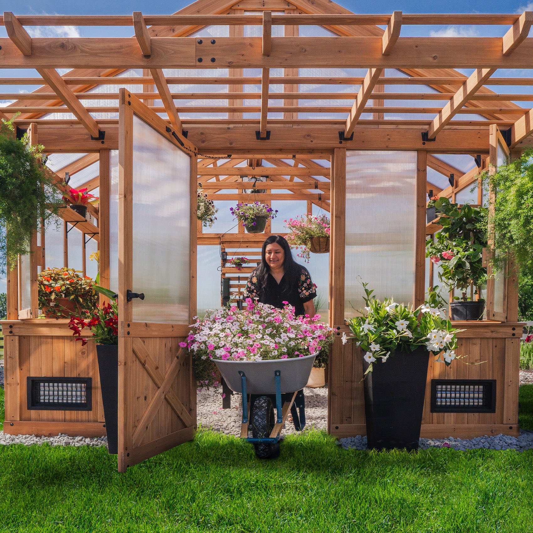 Woman rolling a wheelbarrow of flowers through the wide entry door of the Bellerose XL greenhouse kit.