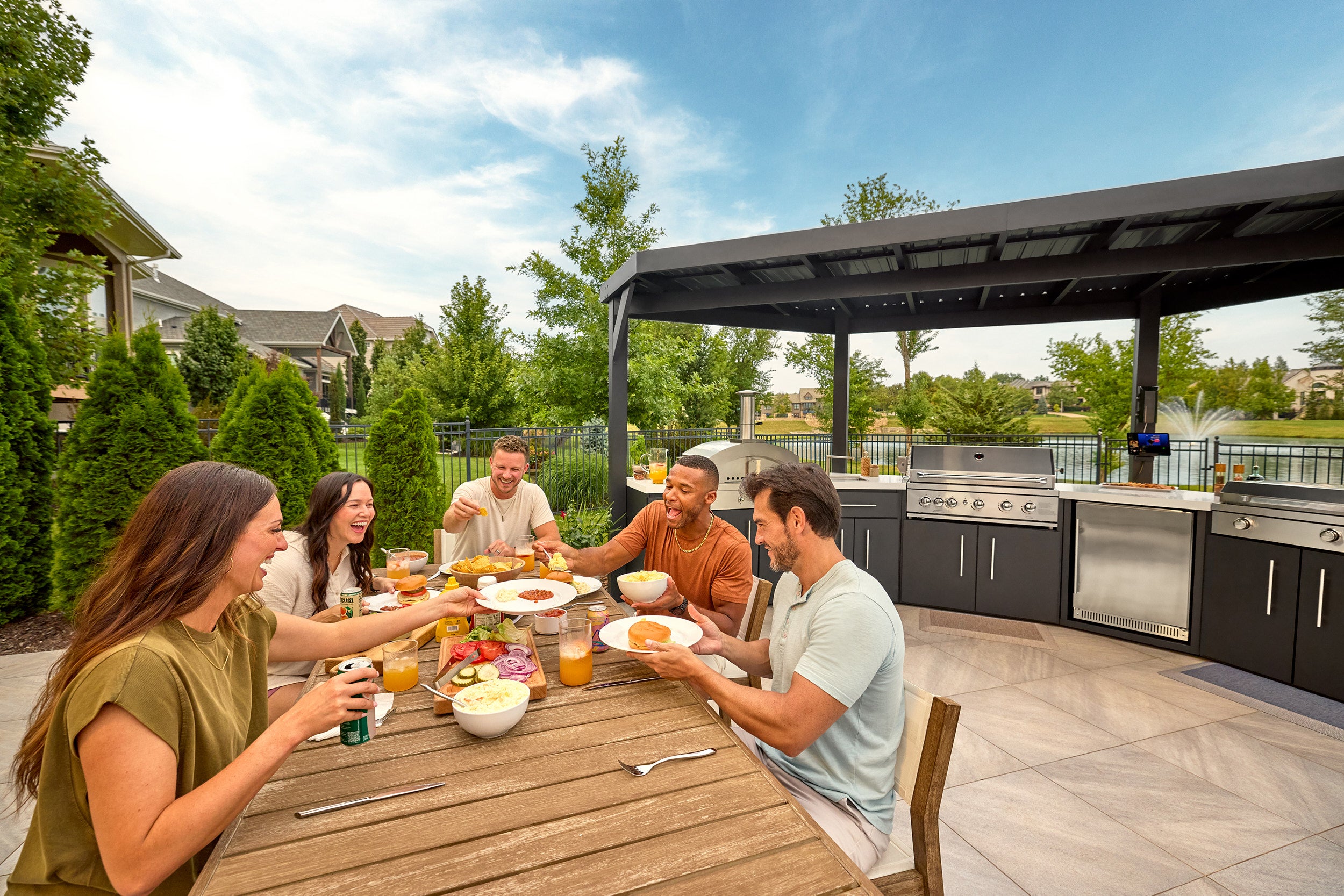 Edgemont Cove Outdoor Kitchen full view showing curved dark charcoal cabinet layout with integrated pizza oven, gas grill, and griddle under a covered pergola on a patio with lakefront backdrop