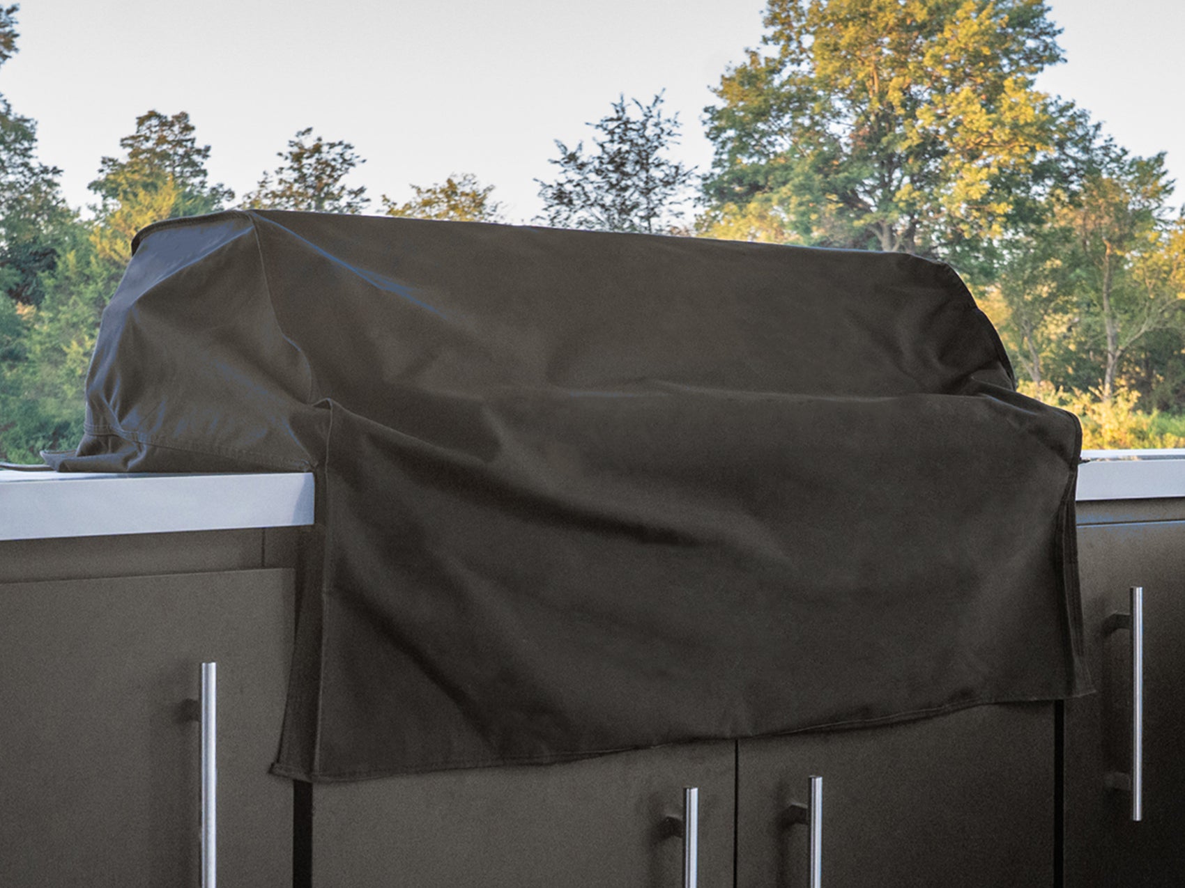 Black protective cover draped over a gas grill in an Emory outdoor kitchen with stainless steel cabinets and fall foliage.