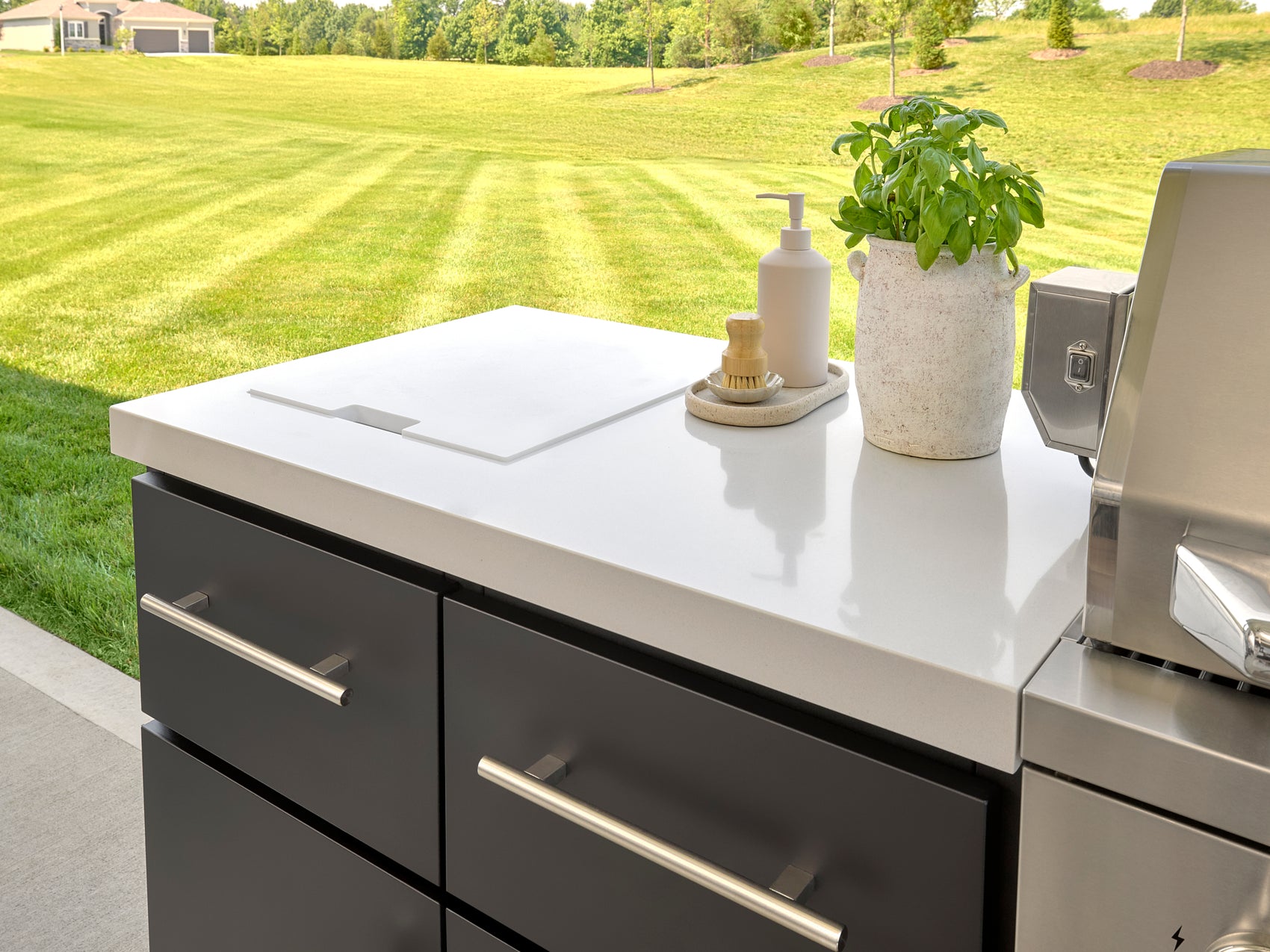 Range Outdoor Living kitchen island with built-in grill and refrigerator on covered patio under steel roof and lawn view.