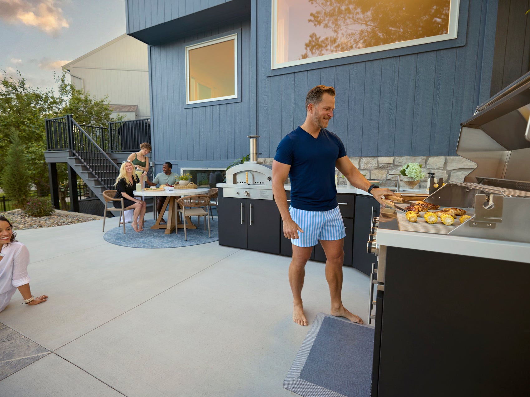 Man grilling at Emory outdoor kitchen with stainless grill, pizza oven, and countertops during backyard gathering.