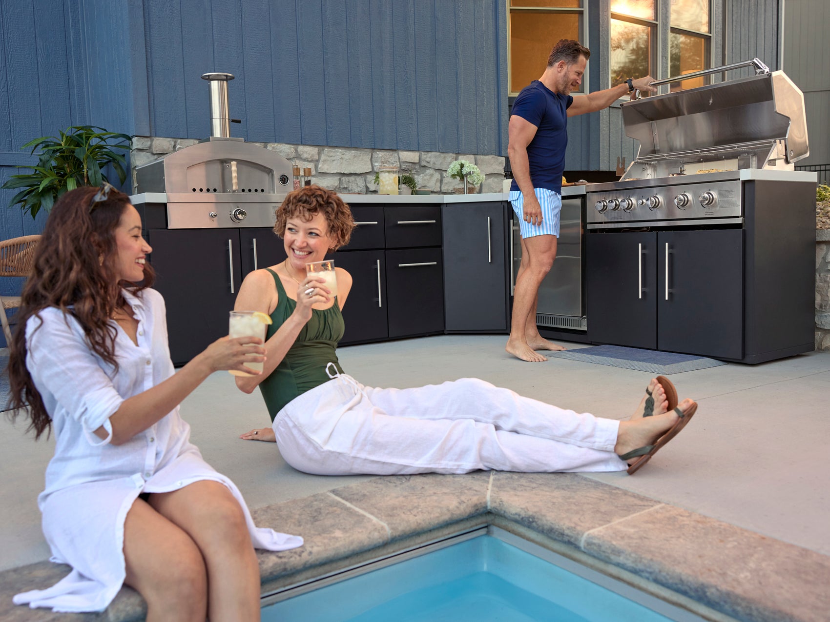 Three people Emory enjoying outdoor kitchen with cabinetry, built-in grill, and pizza oven beside small pool on sunny patio.