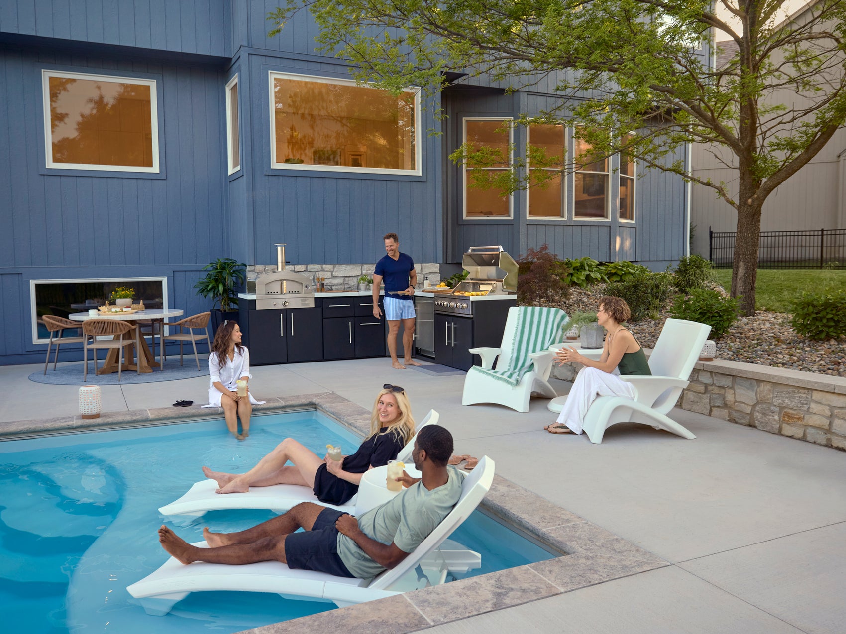 Modern backyard patio with Emory outdoor kitchen, lounge chairs, and contemporary blue home exterior with large windows.