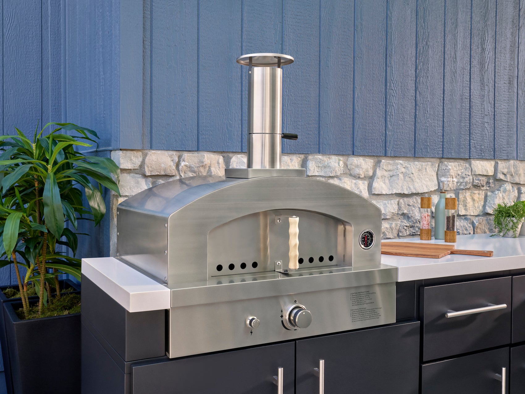 Stainless steel wood fired pizza oven with dome design and chimney built Emory into outdoor kitchen with black cabinets.