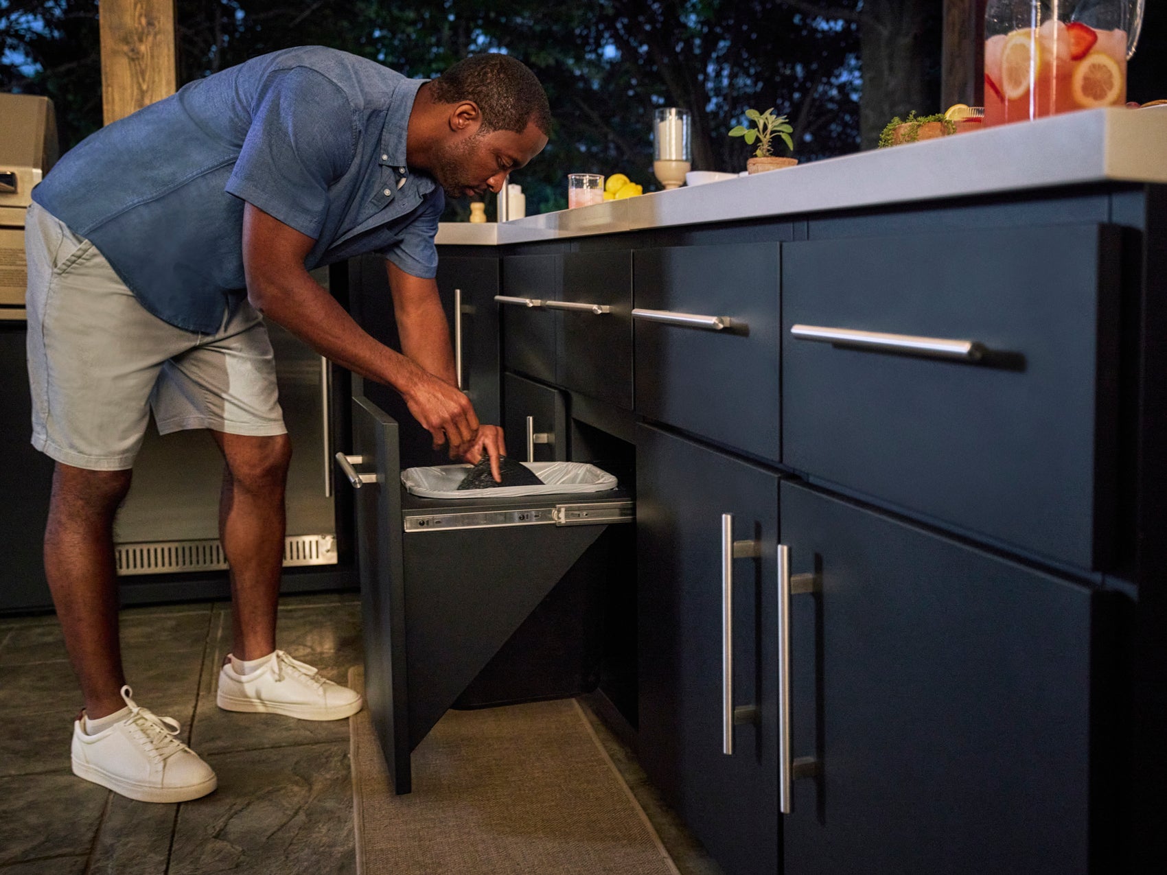 Man disposing waste in pull-out trash bin drawer of Emory outdoor kitchen unit on deck with stone tiles and greenery.
