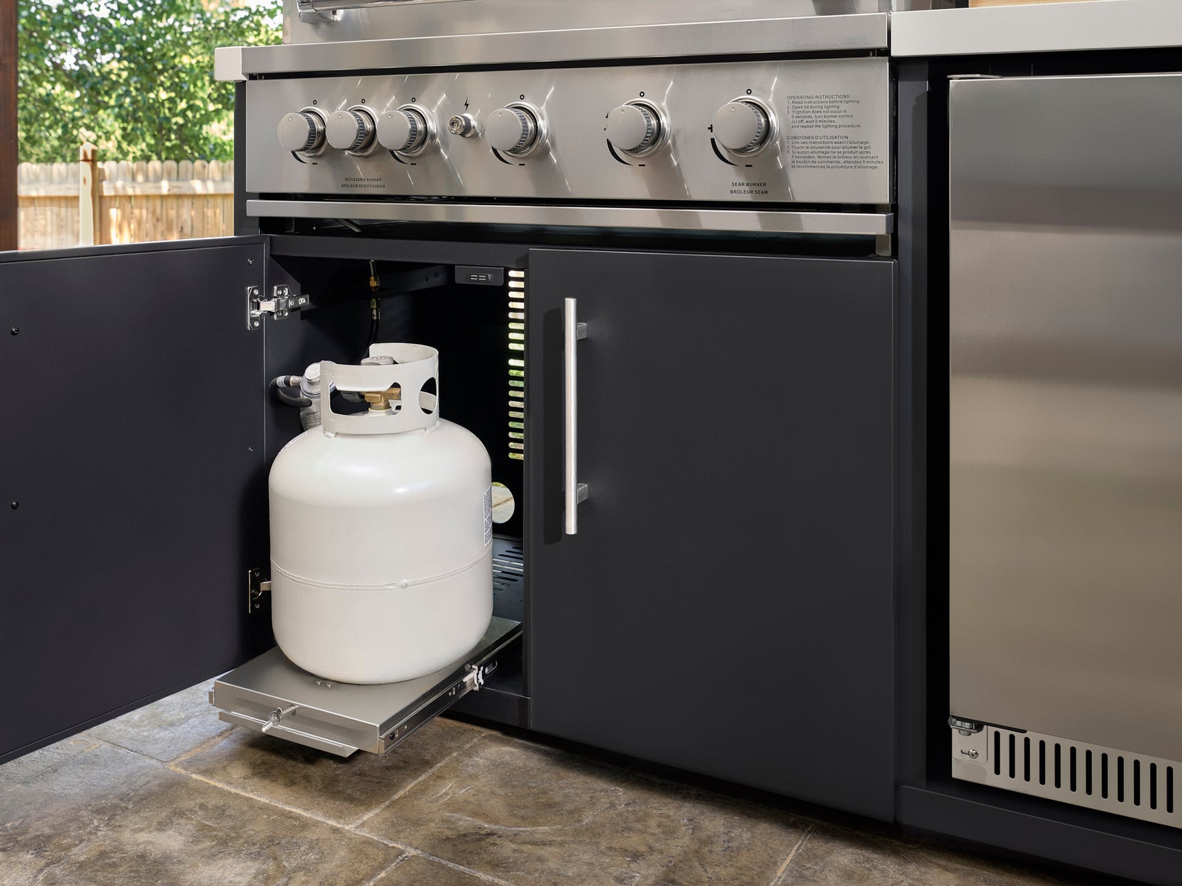 Emory Outdoor kitchen cabinet with propane tank on slide-out tray beneath stainless steel grill with control knobs.