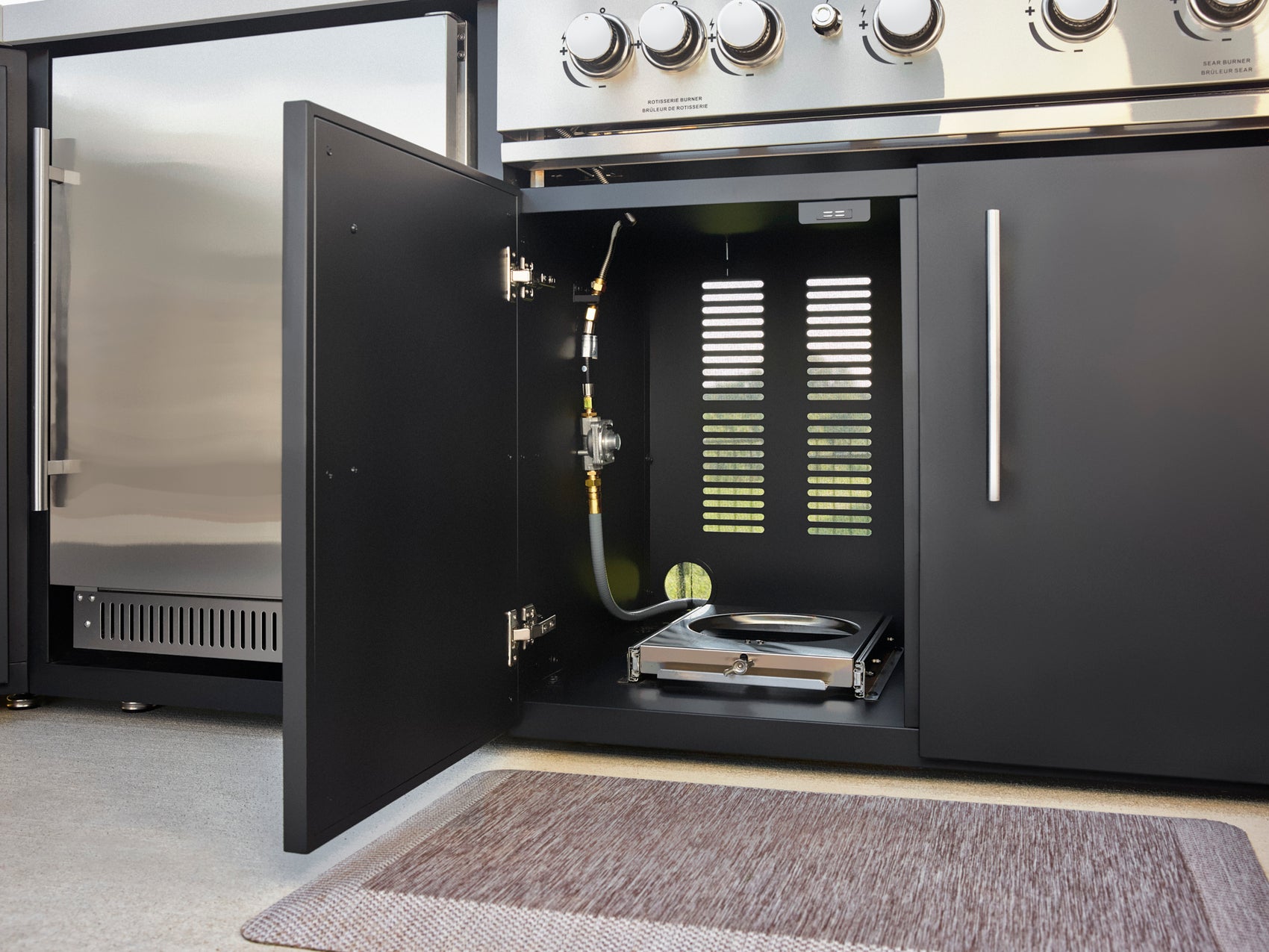 Built-in outdoor gas grill storage compartment with gas line, drip tray, and ventilation slots in matte black cabinet.