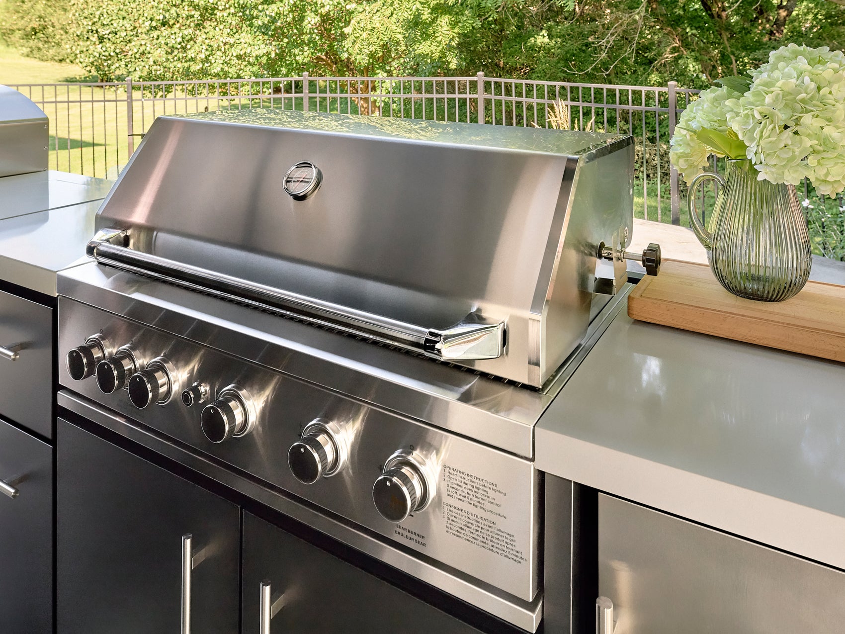Stainless steel built-in grill with control knobs on outdoor bbq island with engineered stone countertop and flowers.