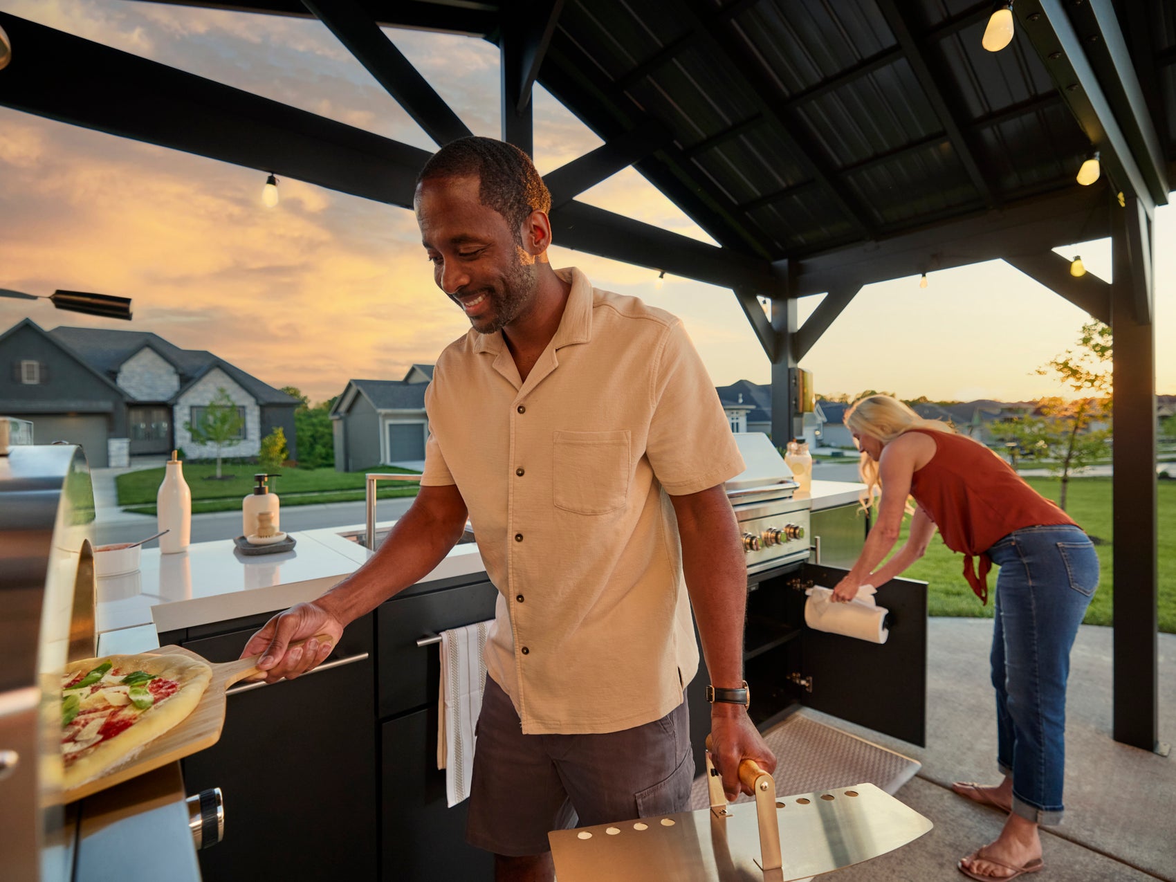 Steel L-shaped outdoor kitchen with built-in grill under peak roof glowing at sunset