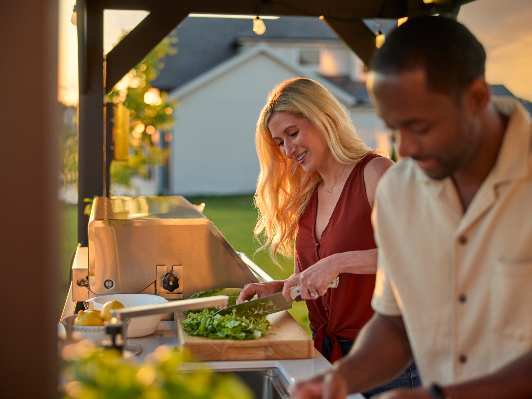 Couple preparing food at covered backyard kitchen with built-in stainless steel grill at golden hour