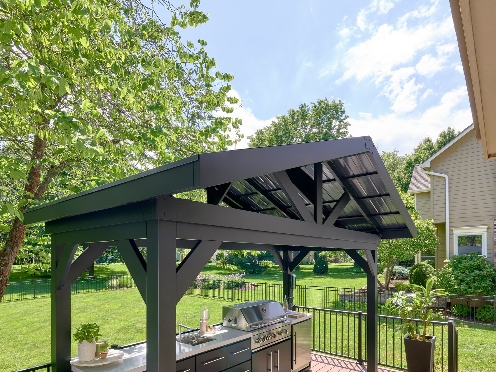 Range Outdoor Living Lancaster outdoor kitchen with steel roof and premium grill in landscaped backyard setting.