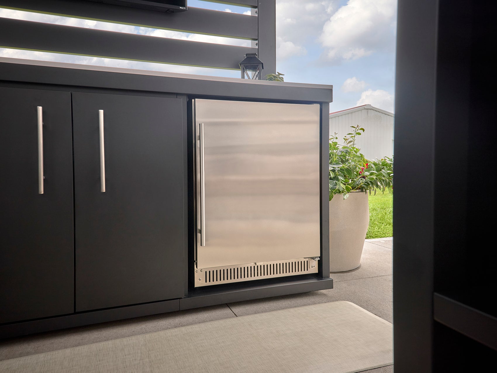 Stainless steel outdoor fridge built into dark gray cabinetry with long bar handles on covered patio with grass yard visible.