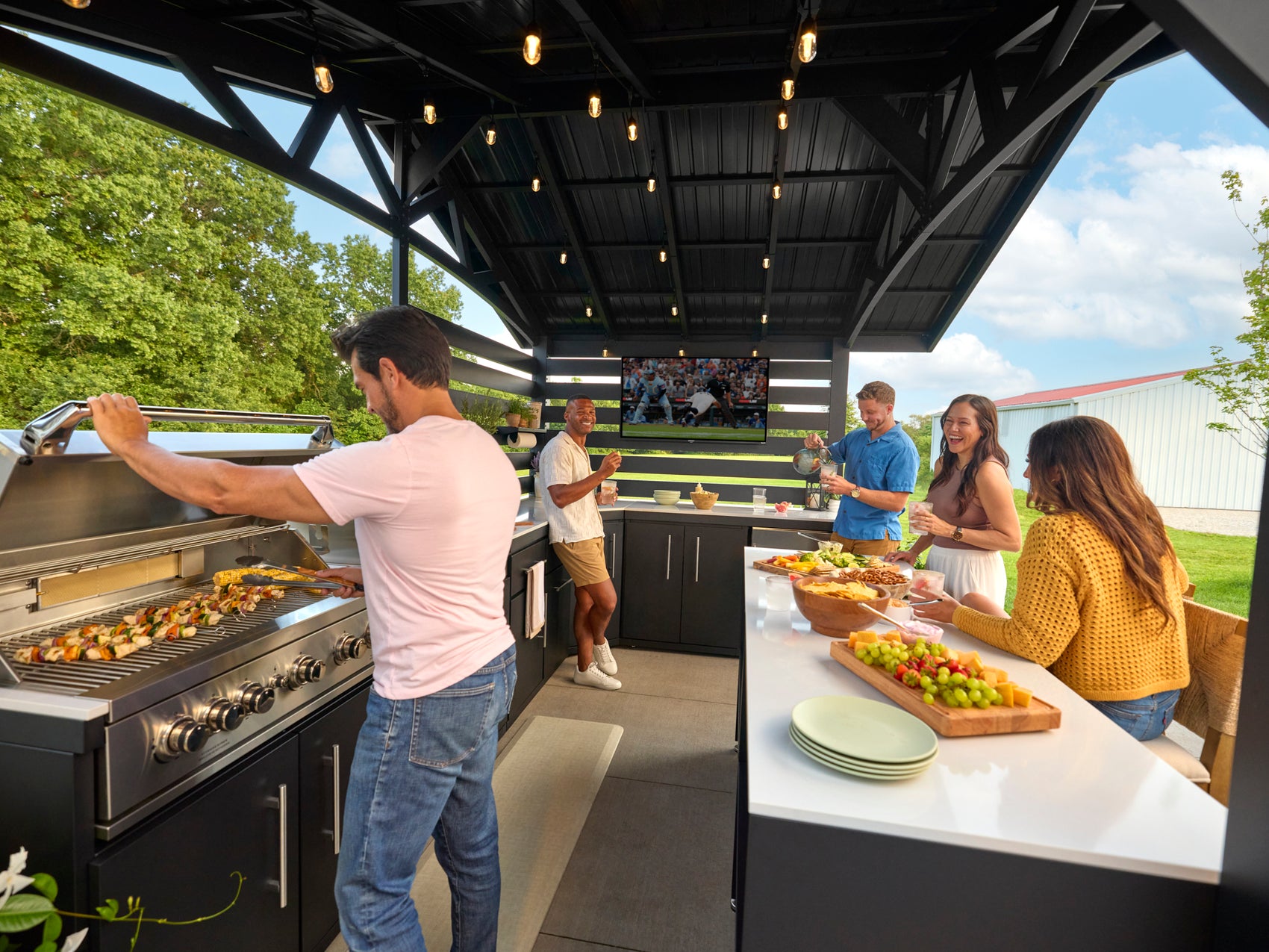 Langston covered outdoor kitchen grill station with people grilling and socializing under black pavilion and TV.