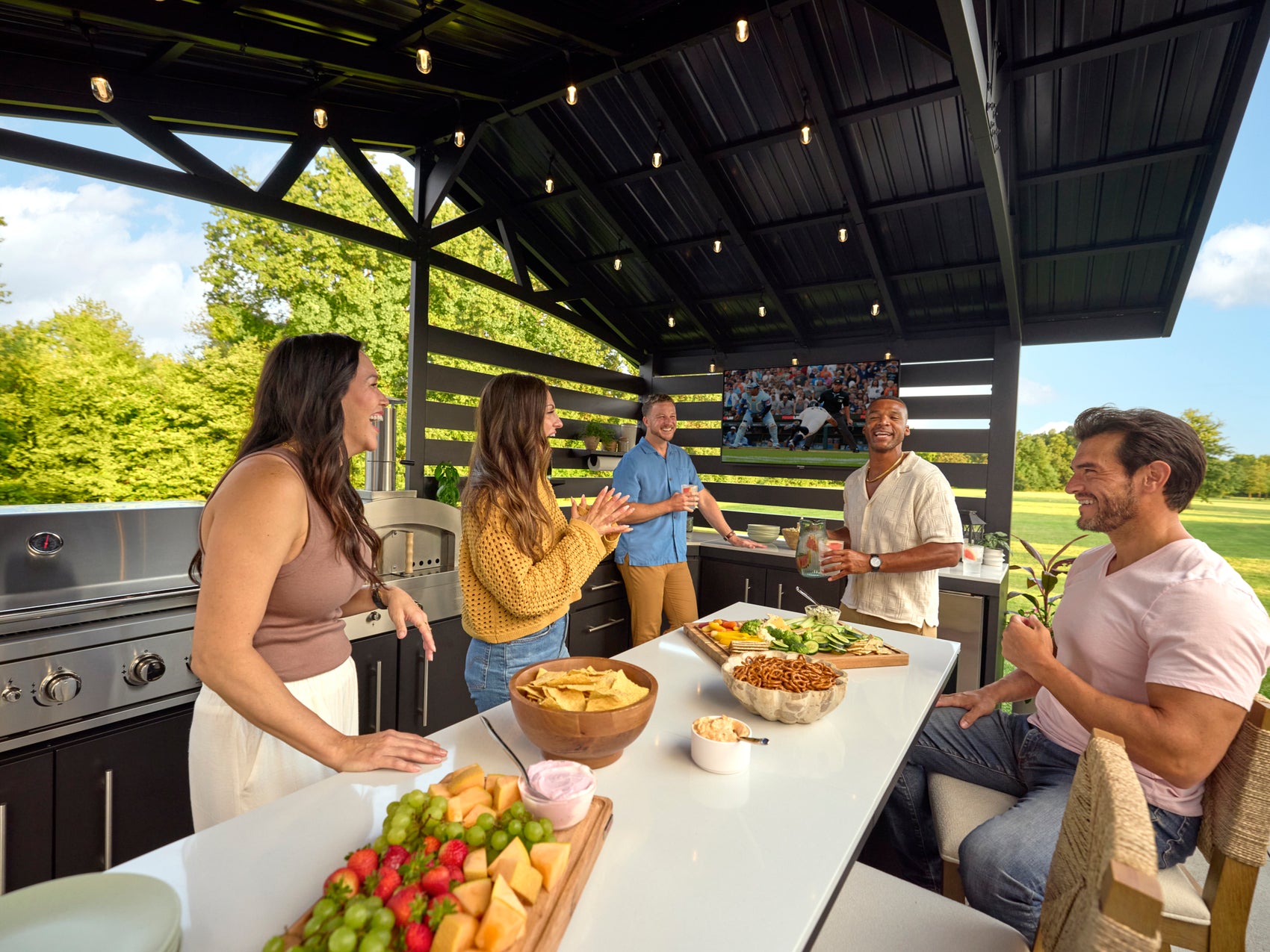 Friends gathered around an Langston outdoor kitchen island with grill, TV, and countertop under covered patio.