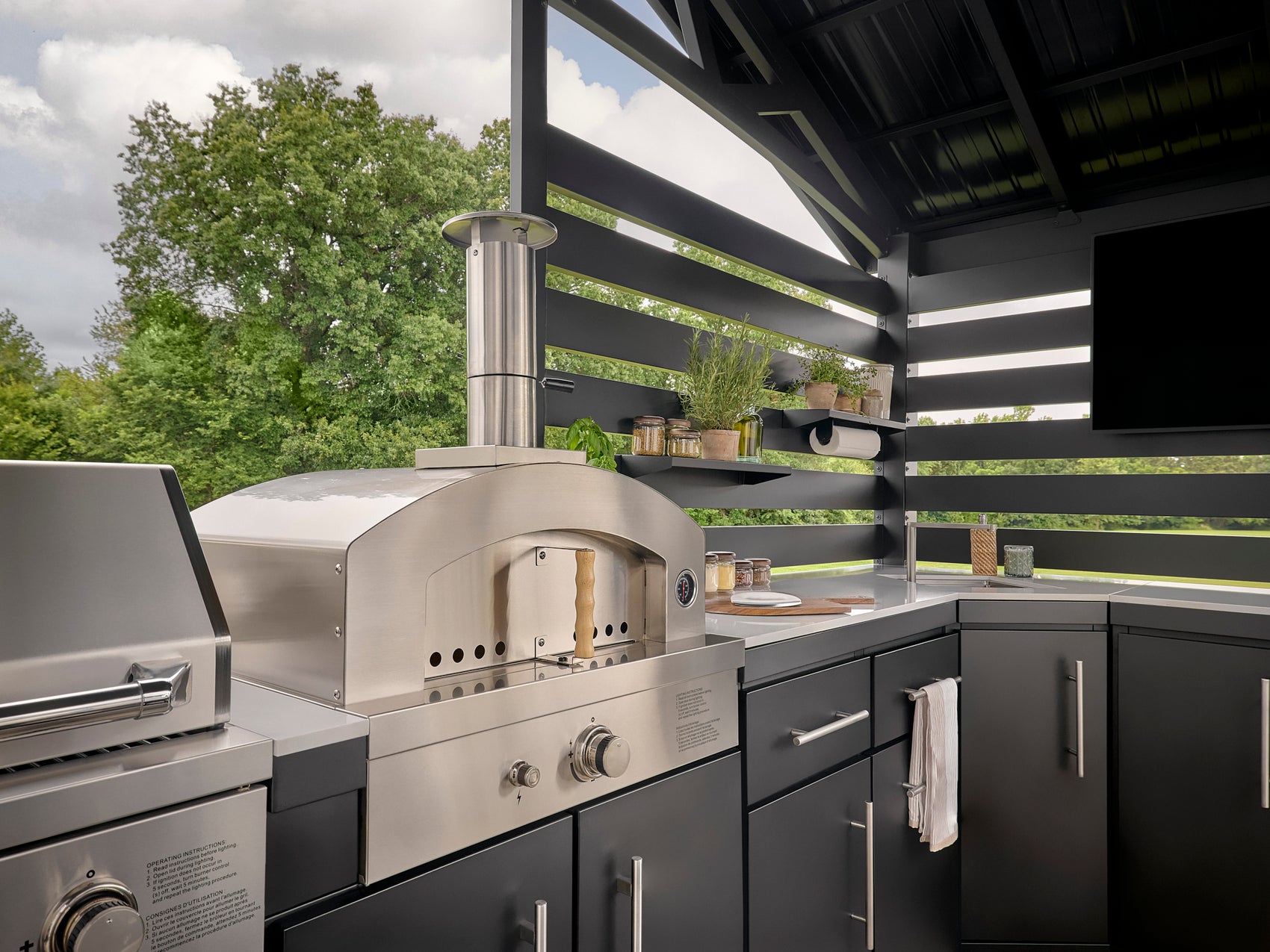 Stainless steel outdoor pizza oven with arched opening built into dark gray cabinetry under covered patio.