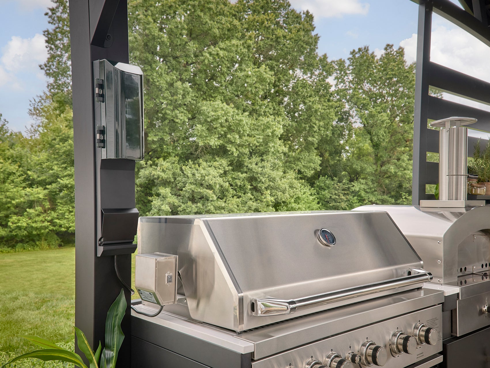 Langston outdoor kitchen with a built-in grill and a TV mounted on a black steel roof beam, with green trees behind.