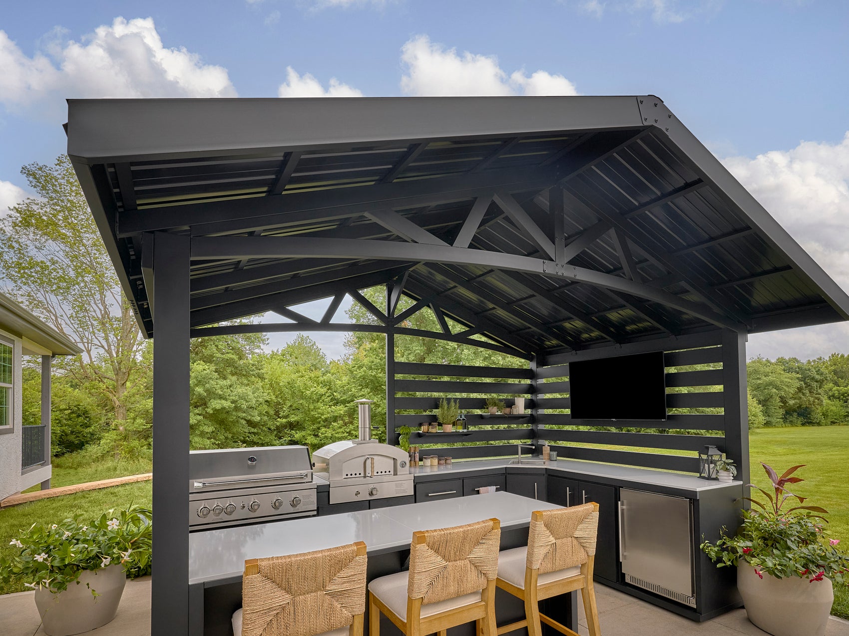 Attached steel roof with trellis panels over the Langston outdoor kitchen with a grill, bar seating, and a mounted TV.