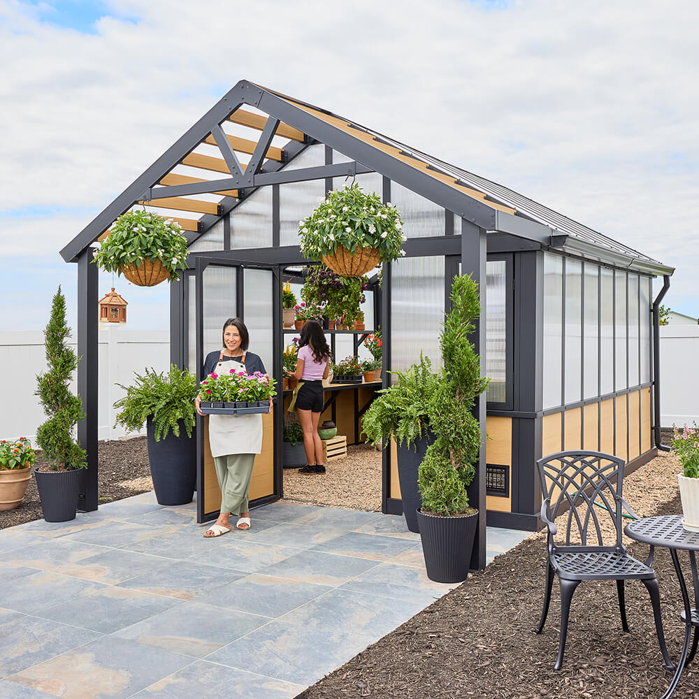 Lifestyle image of women inviting you into your new Larkspur metal and composite wood greenhouse. Click image to shop the Greenhouse category.