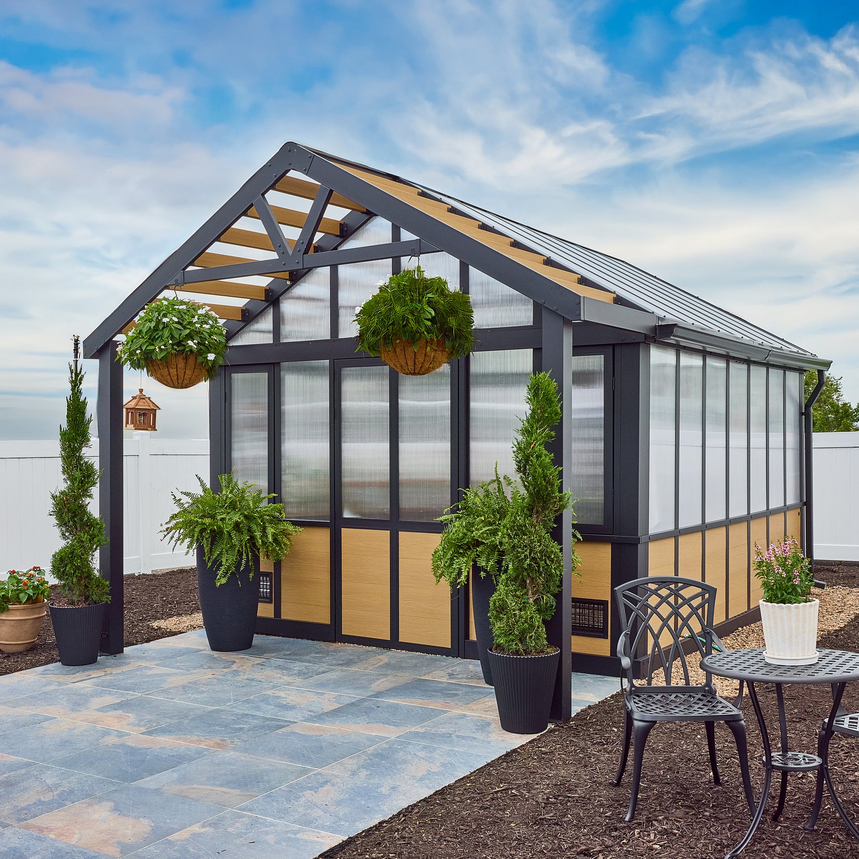 The Backyard Discovery Larkspur metal greenhouse with a peaked roof and polycarbonate panels on a paved foundation.