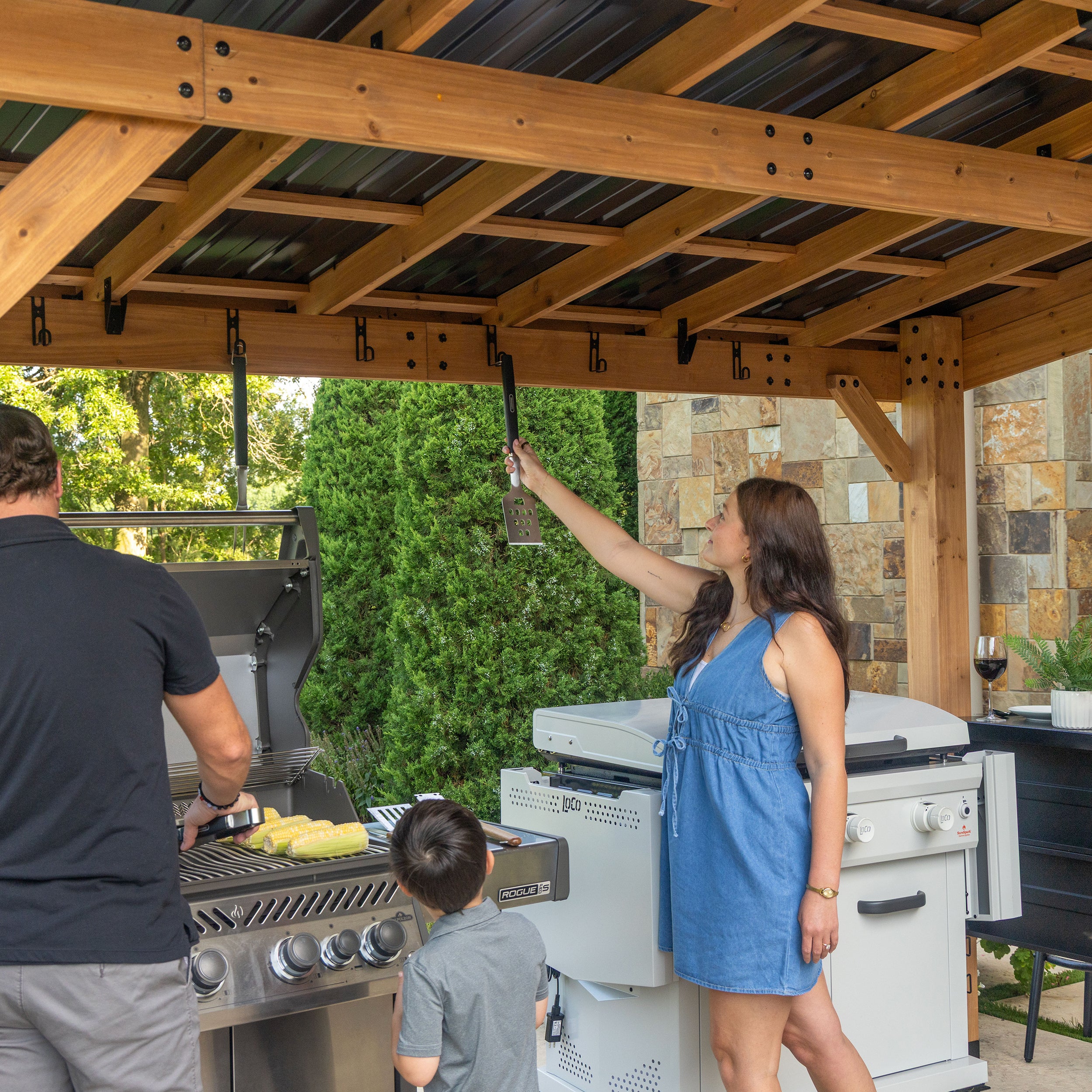 Cedar grill gazebo with accessory hooks, steel roof beams, and outdoor kitchen stone backdrop.