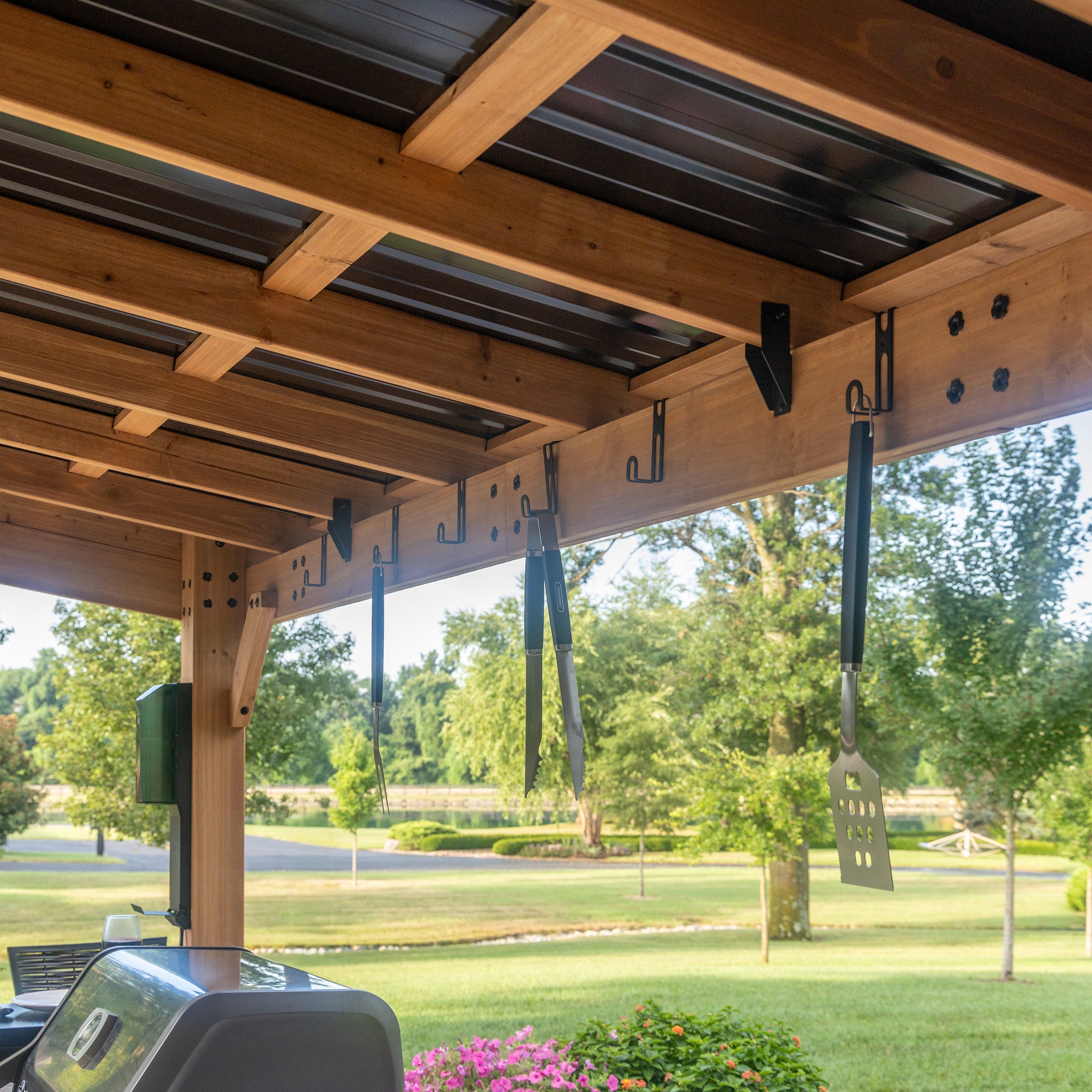 Steel accessory hooks mounted on cedar beam of metal grill gazebo overlooking backyard lawn.