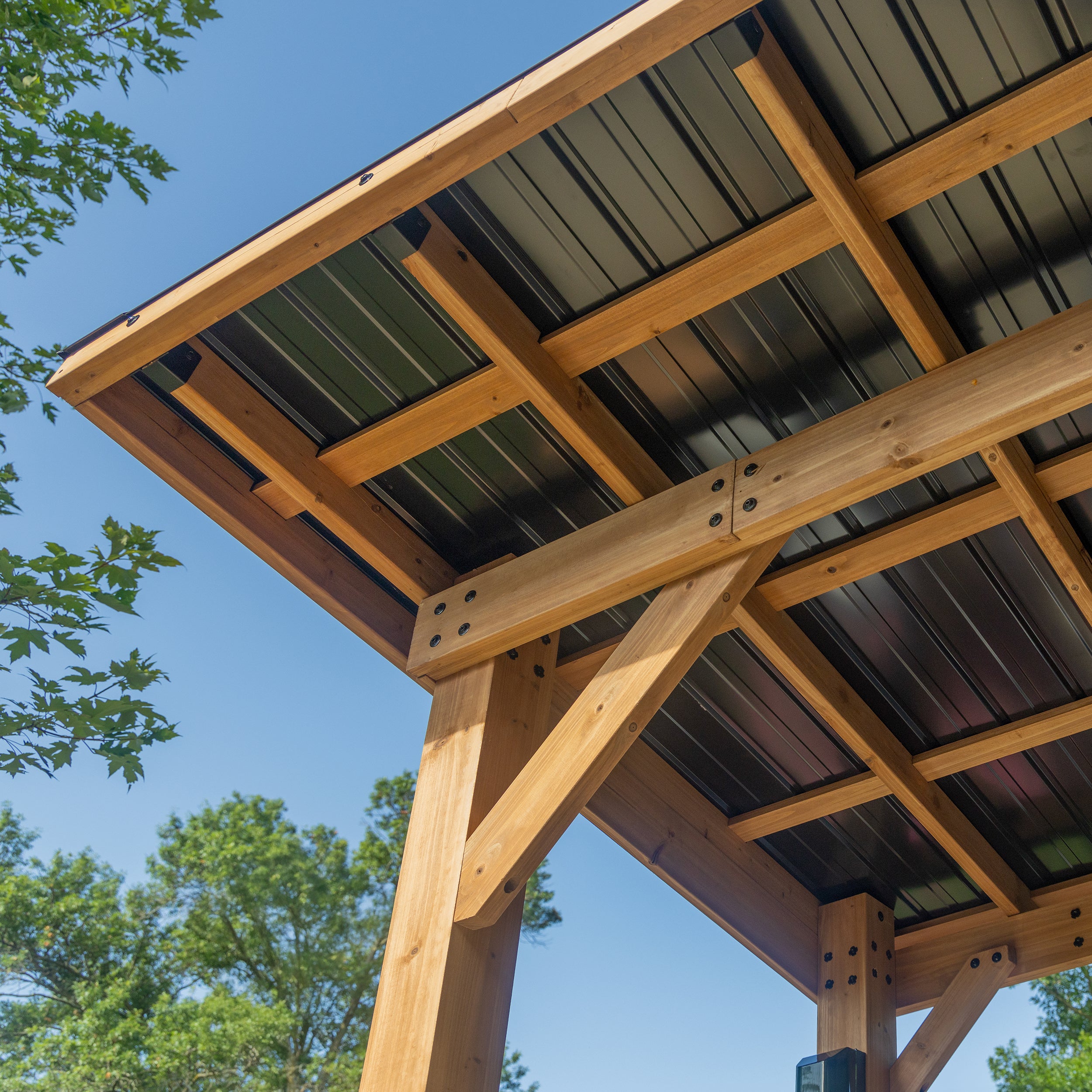 Underside view of metal roof grill gazebo showing cedar beam frame and steel panels against blue sky.