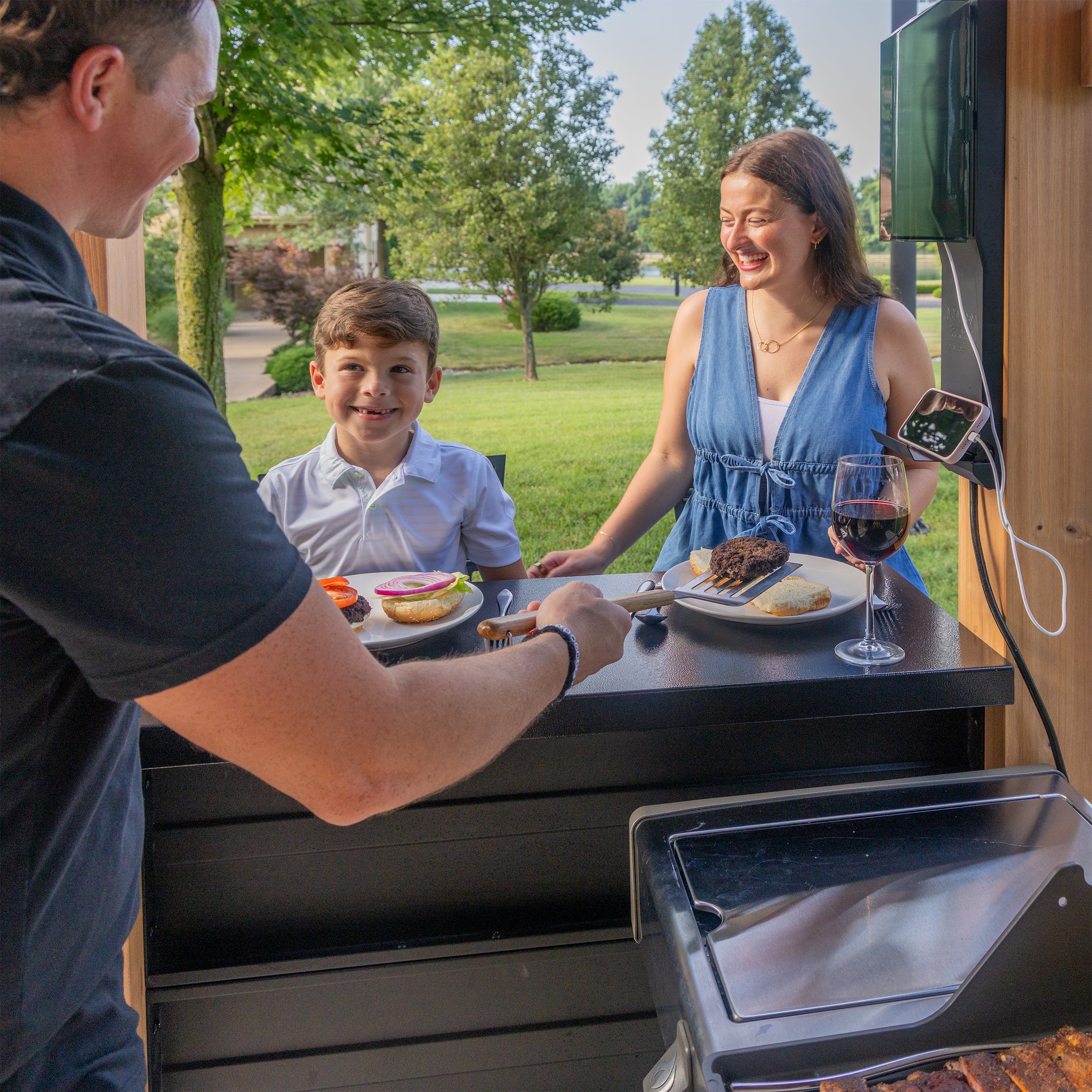 Family enjoying grilled meal at barbecue gazebo with steel countertops in backyard setting.
