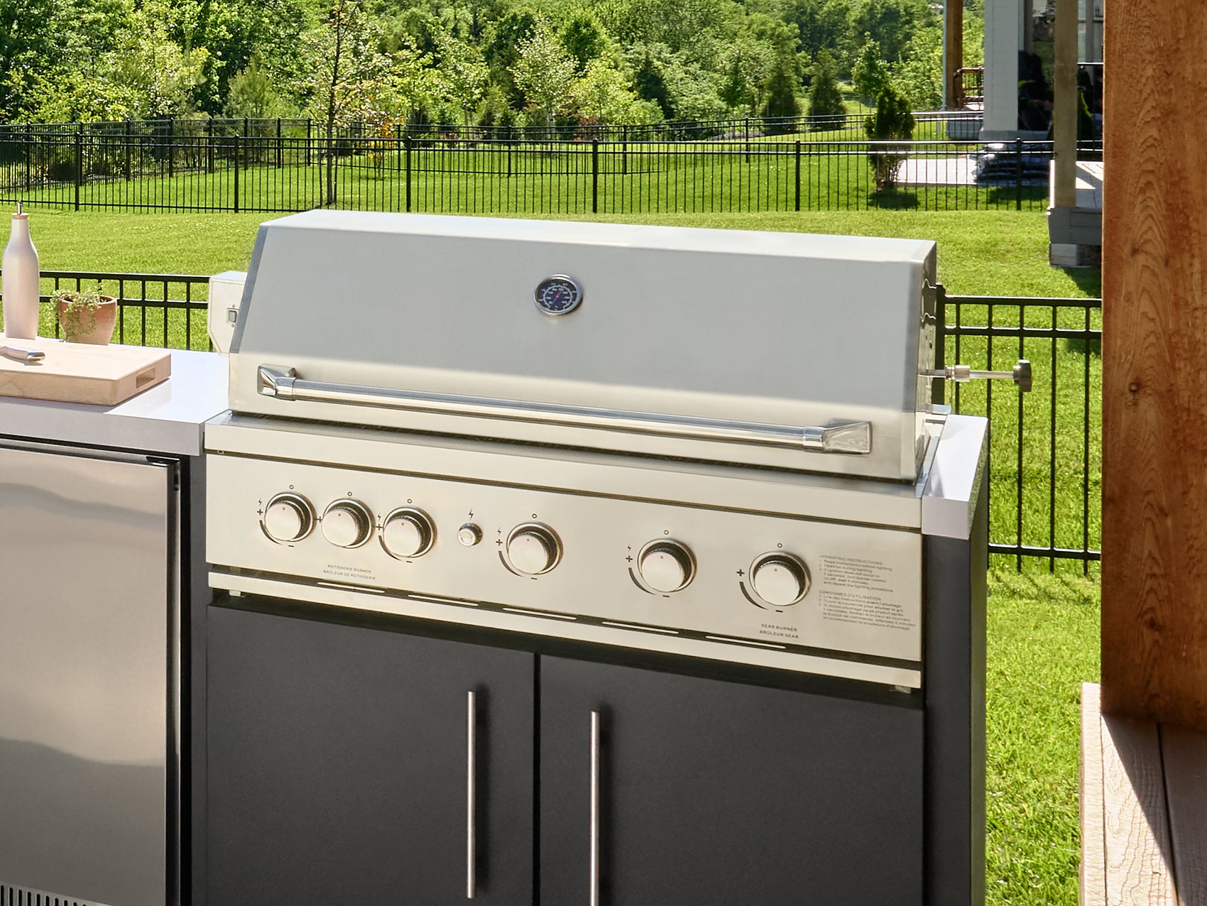 Stainless steel built-in grill with closed lid and temperature gauge, with six control knobs on outdoor patio.