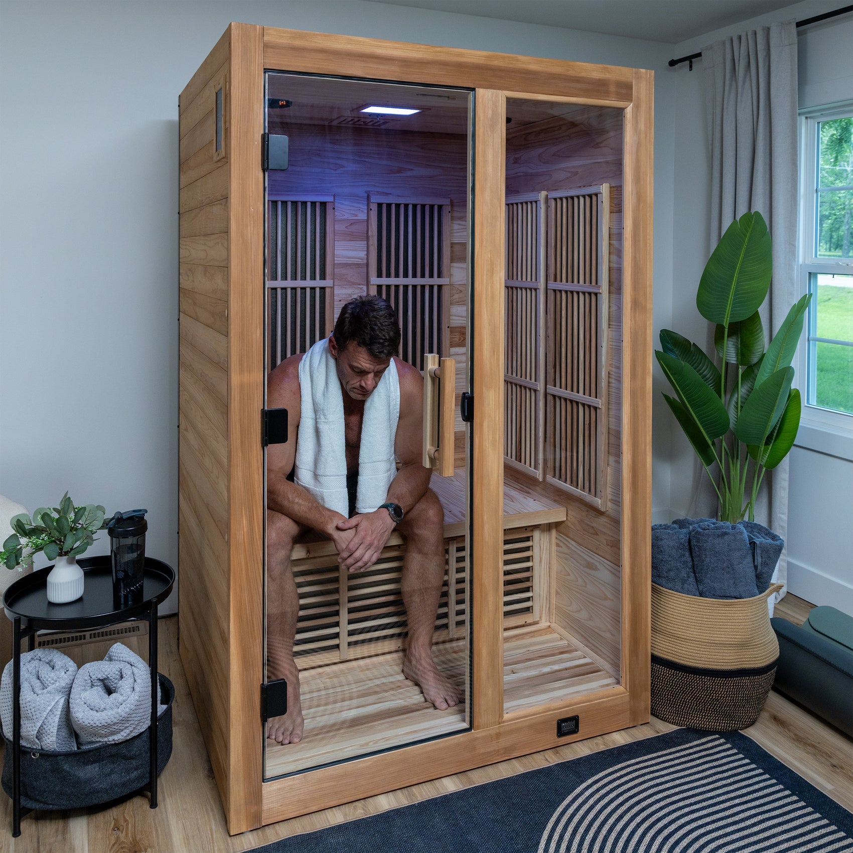 Man sitting inside a wooden infrared sauna with towels and plants around.