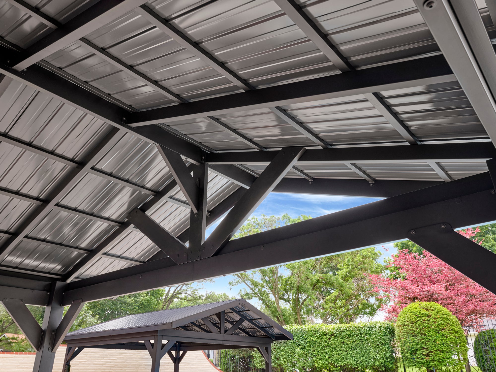Steel backyard gazebo with corrugated metal roof panels and exposed black beam framework viewed from below against blue sky.