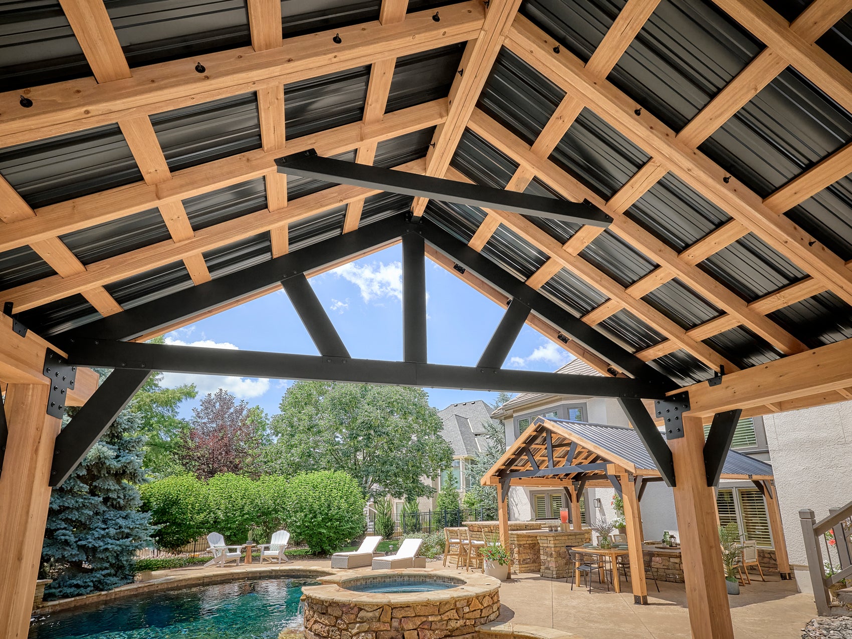 Lodge steel gazebo with metal roof and exposed timber beams over backyard pool, hot tub, and outdoor kitchen area.