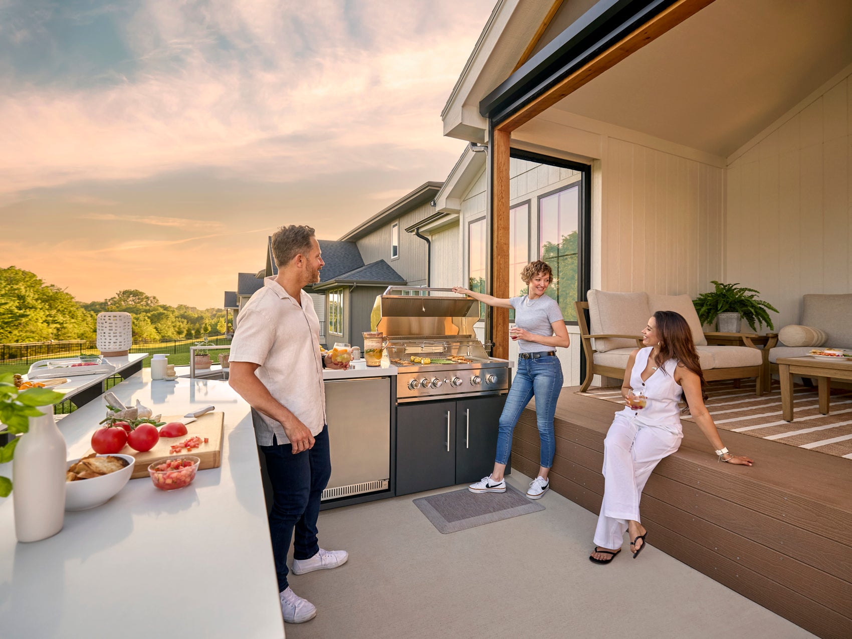 Three people enjoying outdoor gathering on patio with steel pergola beam overhead, built-in grill, and modern furnishings.
