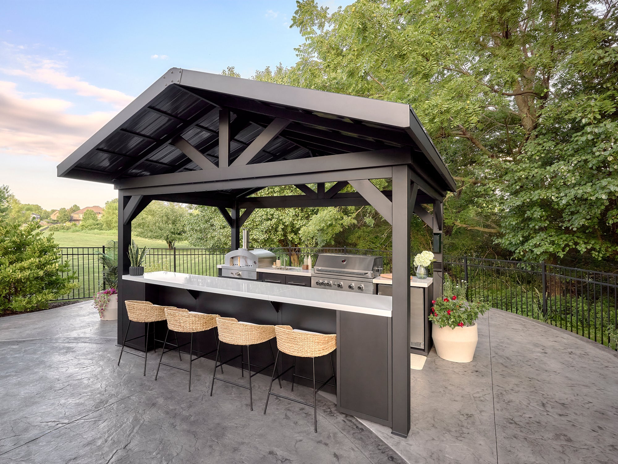 Range Outdoor Living Lancaster galley kitchen with black pavilion roof, engineered stone bar, grill and pizza oven on patio.