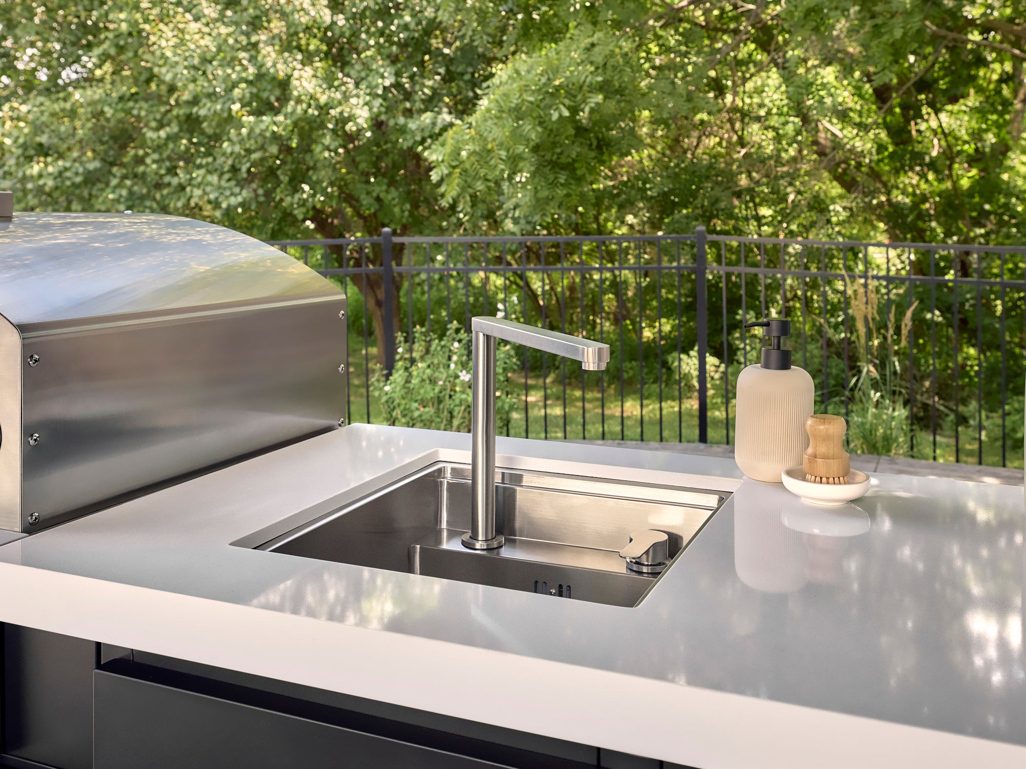 Stainless steel sink with modern faucet set into white countertop on Lancaster outdoor kitchen with grill and greenery.