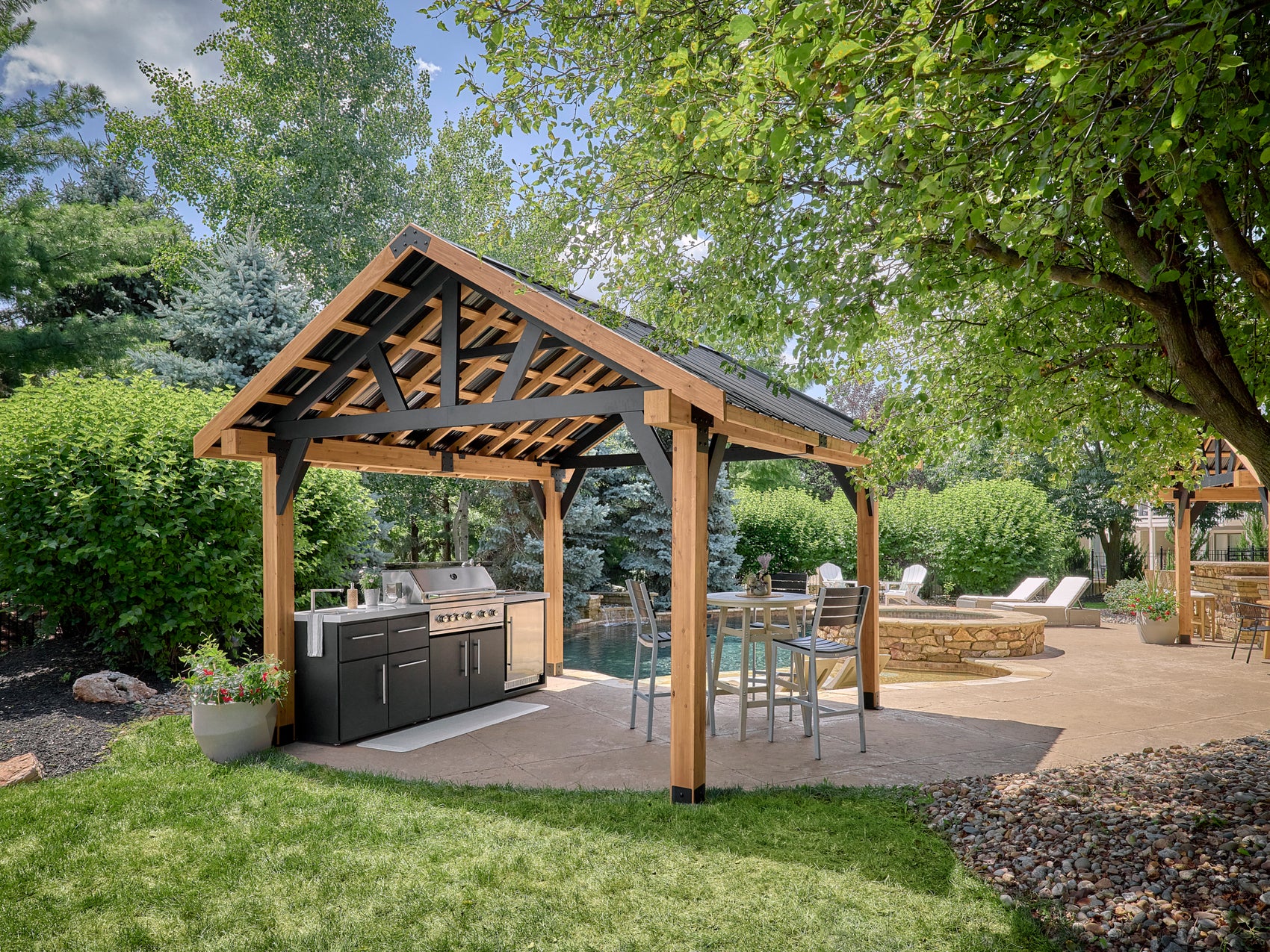 Steel pavilion gazebo with exposed wood-look beams creating backyard shade over outdoor kitchen, bar seating, and pool patio.