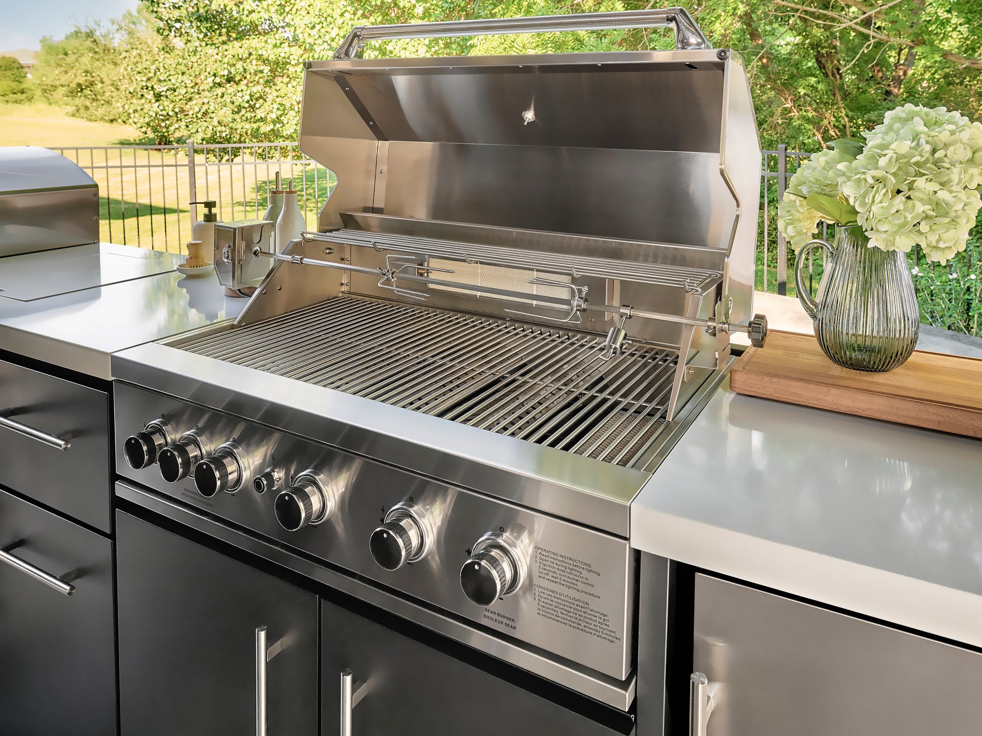 Open stainless steel bbq grill with rotisserie rod and cooking grates on Lancaster outdoor kitchen countertop.