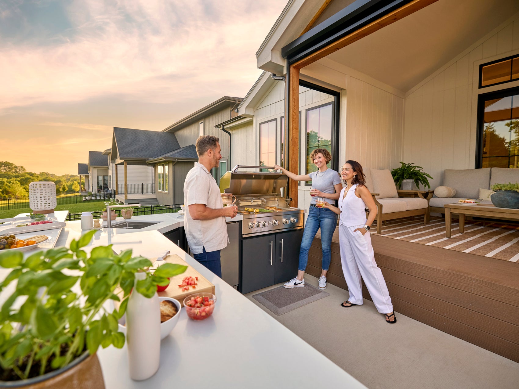 Emory Outdoor kitchen with grill on covered patio where three people gather at sunset with modern white home in background.