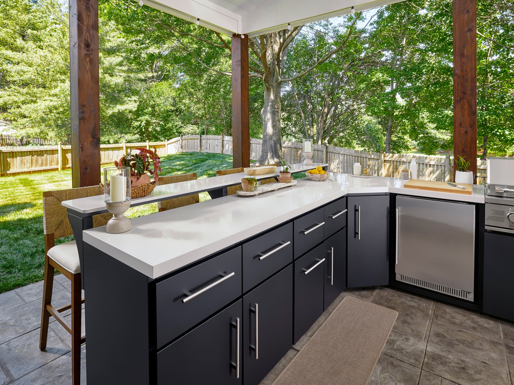 Emory Outdoor kitchen countertop with black cabinetry and bar seating overlooking fenced backyard with mature trees.