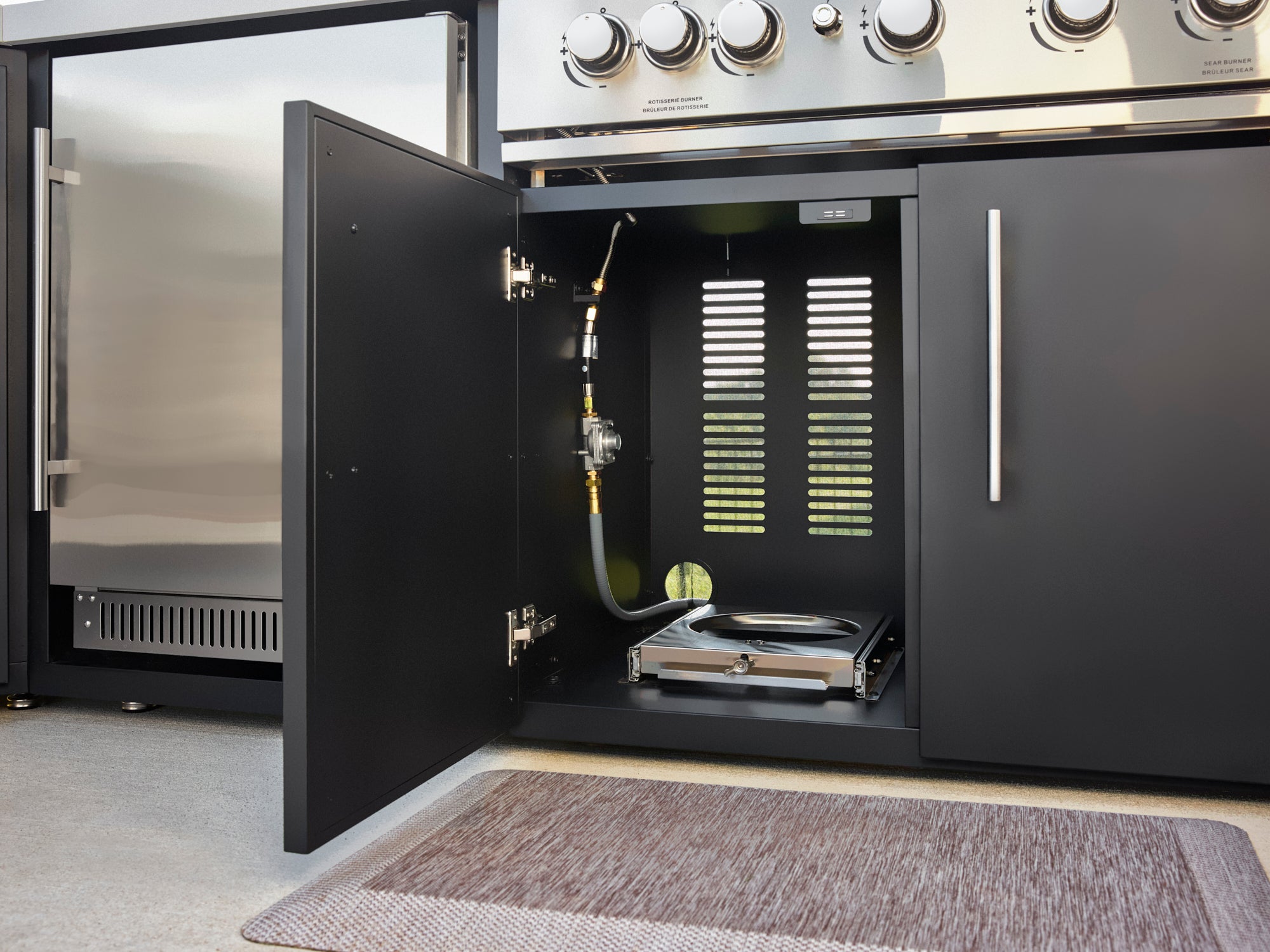 Built-in outdoor gas grill storage compartment with gas line, drip tray, and ventilation slots in matte black cabinet.