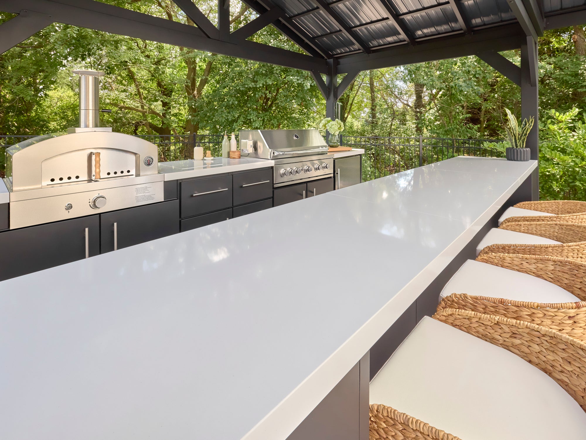 White engineered stone outdoor kitchen counter with bar seating under covered patio with dark cabinetry and appliances.