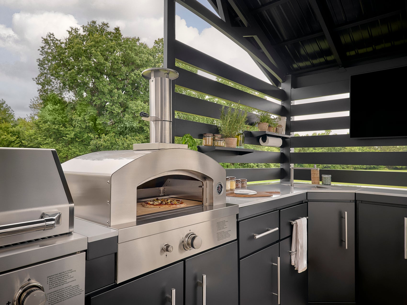 Langston stainless steel outdoor pizza oven with arched door and control knobs built into dark gray cabinetry.