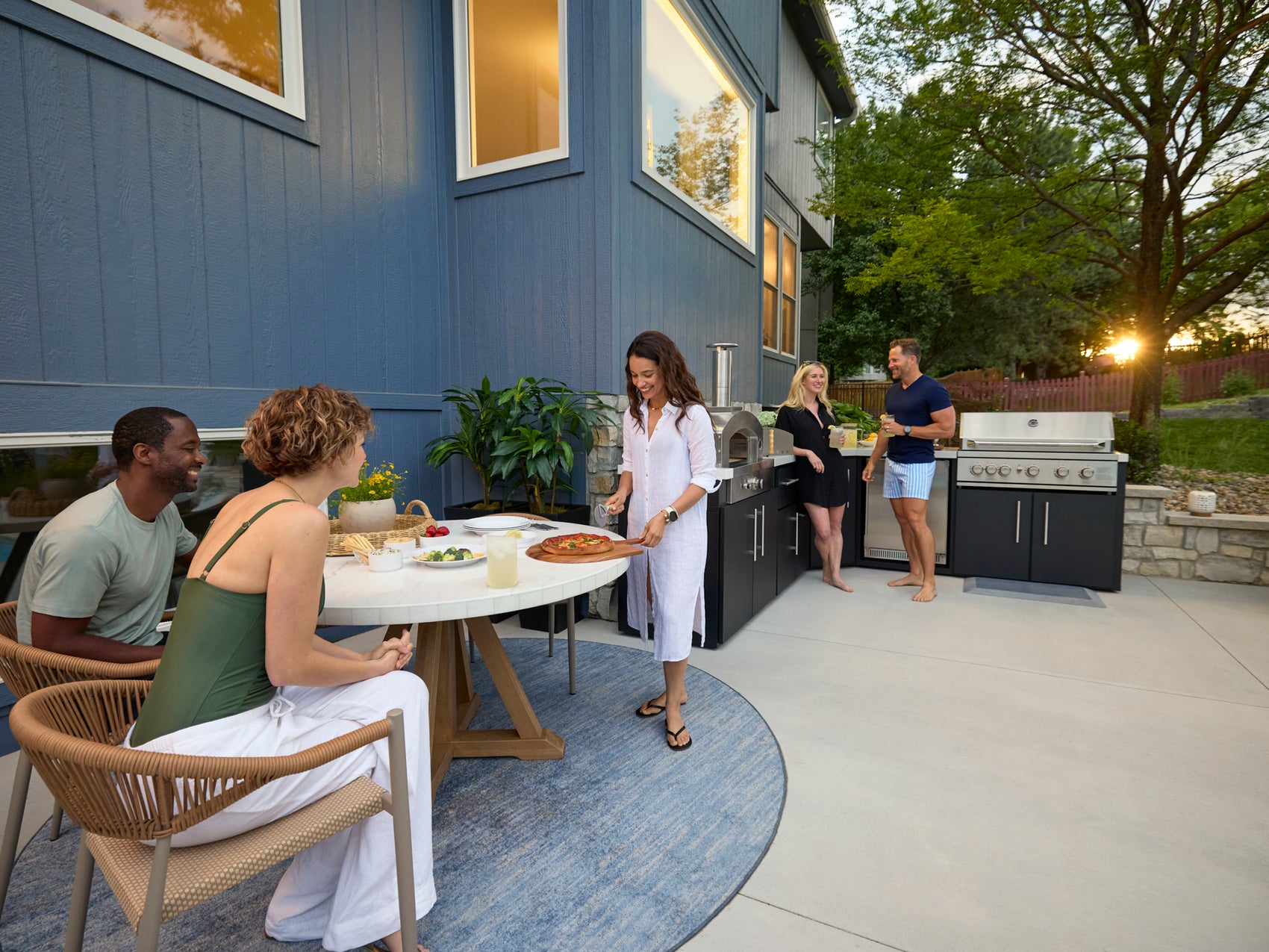 Friends gathered on modern patio with Emory outdoor kitchen, dining area, and attached pergola beside contemporary blue home.