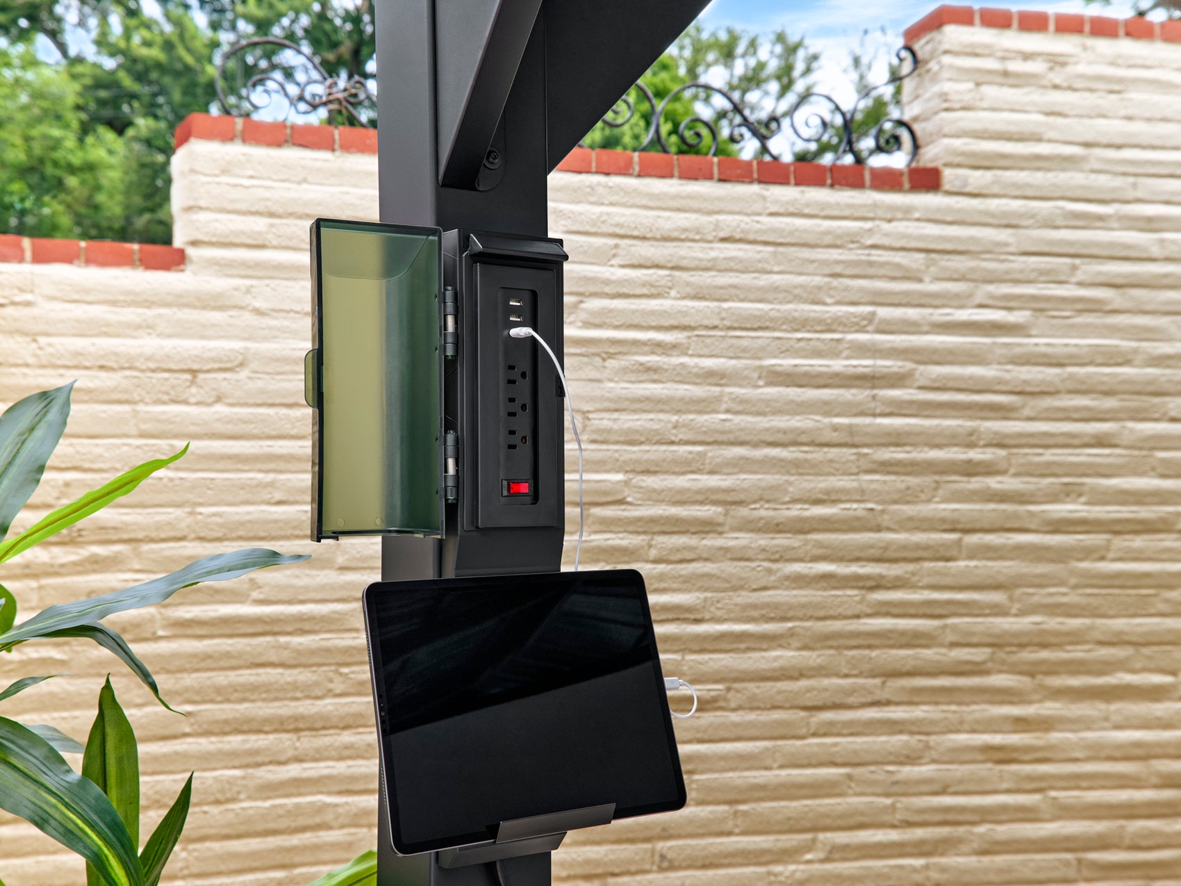 Built-in power outlet with USB ports and tablet holder mounted on steel gazebo post against cream stone wall backdrop.