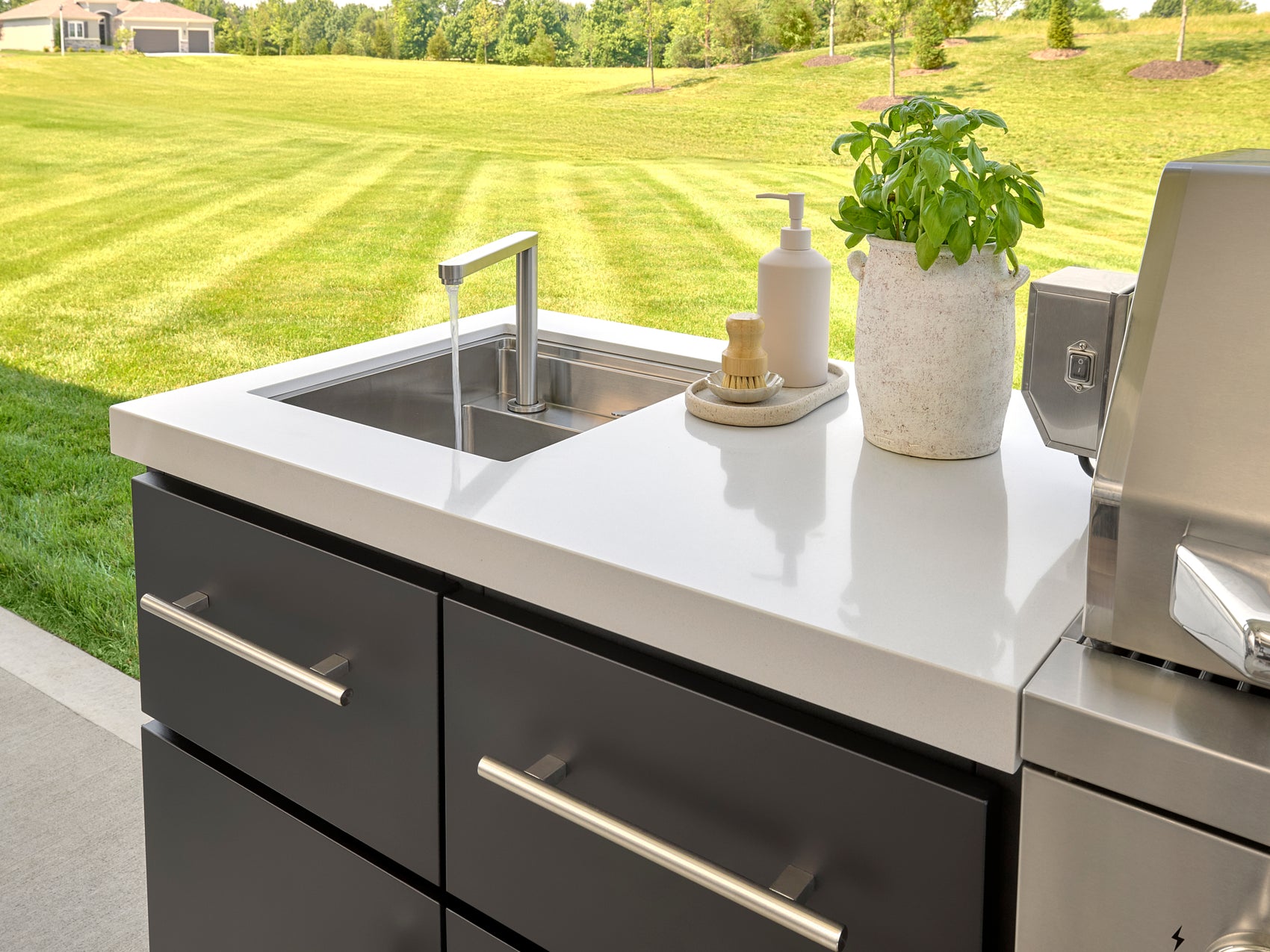 Emory Outdoor kitchen sink with modern faucet set in white engineered stone counter with cabinet drawers and basil plant.